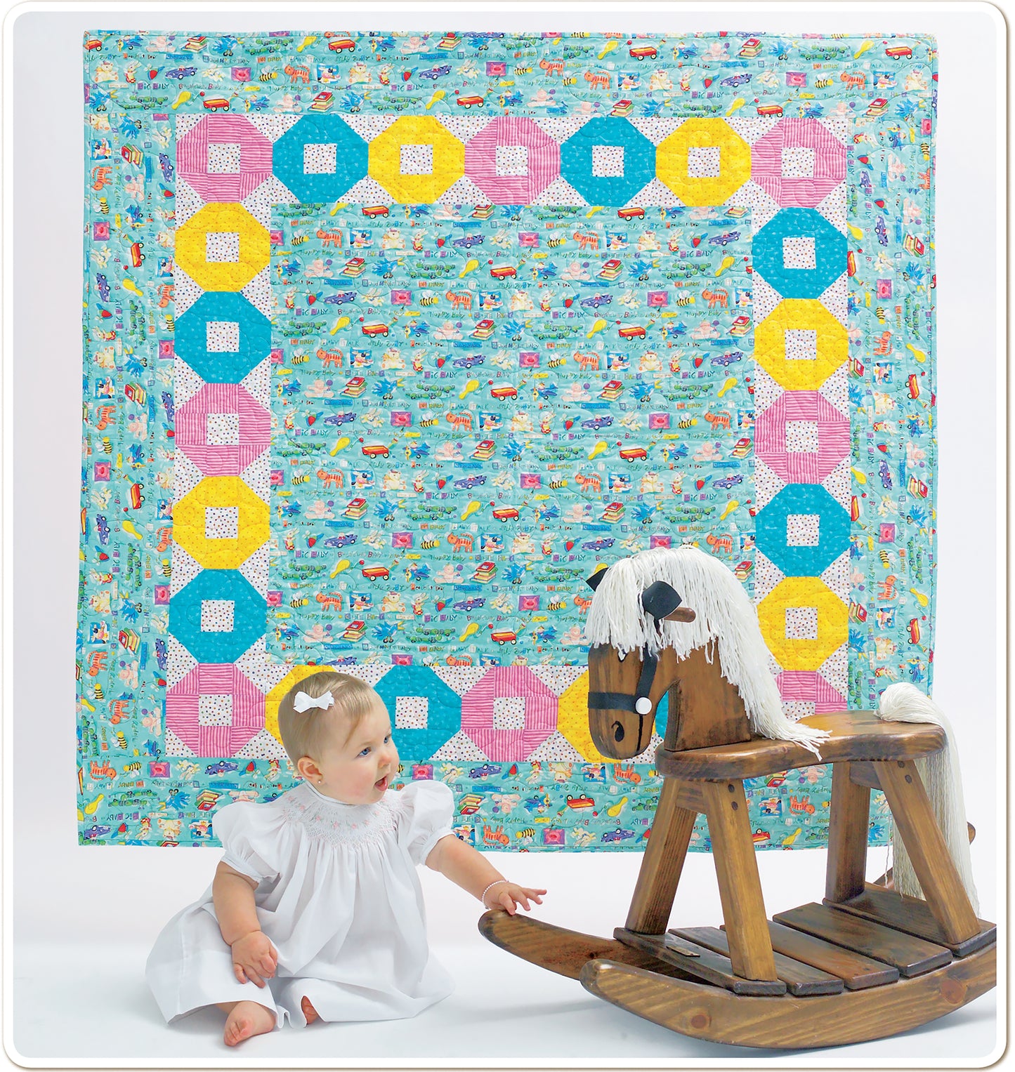 A baby in a white dress sits on the floor reaching for a wooden rocking horse, with a colorful quilt from Leisure Arts’ "Baby Quilts—Pamper Little Ones with Fast Blankets in Fun Fabrics!" hanging in the background.