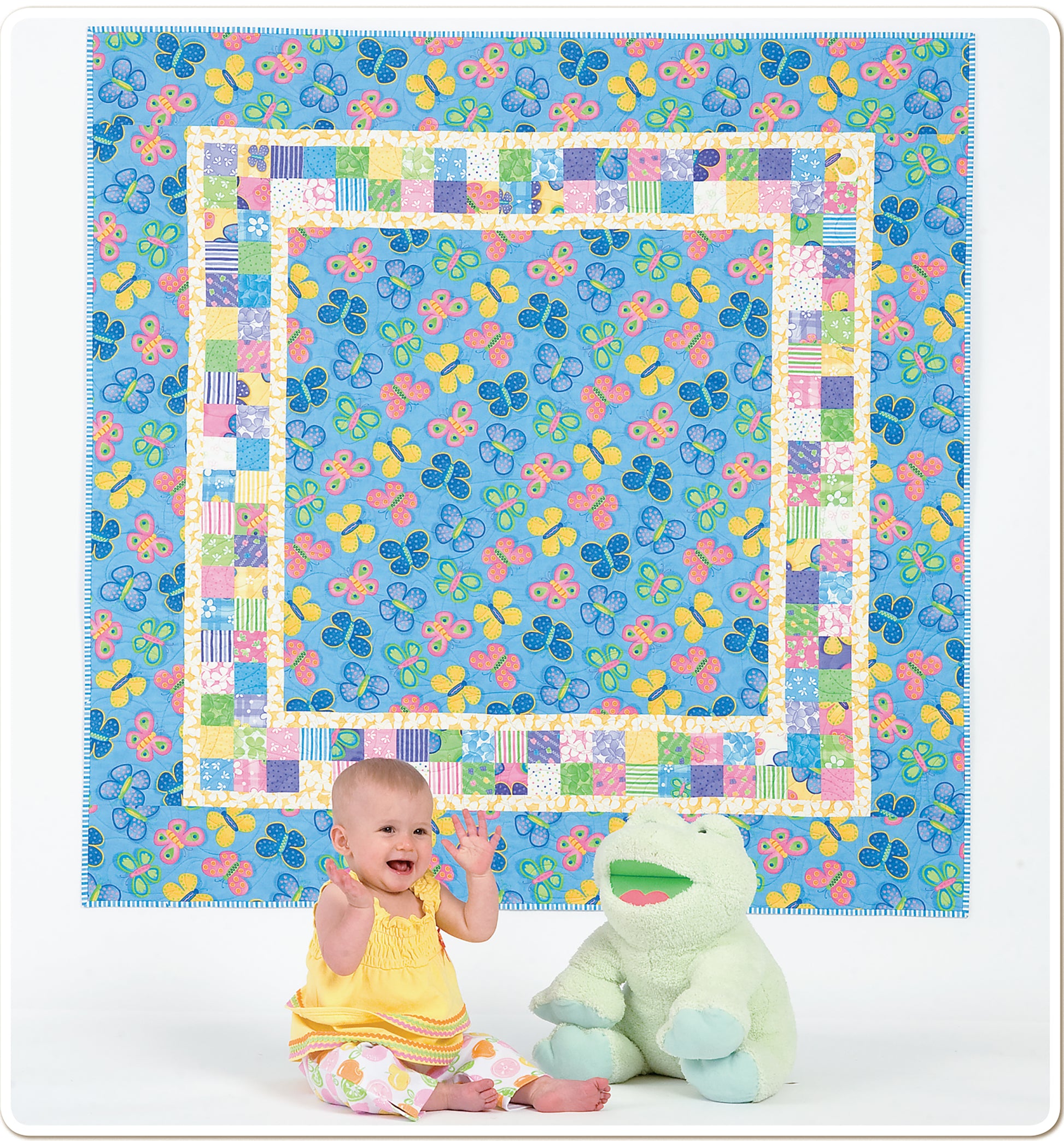 A smiling baby in yellow sits by a large green frog plush toy, in front of a colorful butterfly-patterned quilt from Leisure Arts’ "Baby Quilts—Pamper Little Ones with Fast Blankets in Fun Fabrics!" collection.