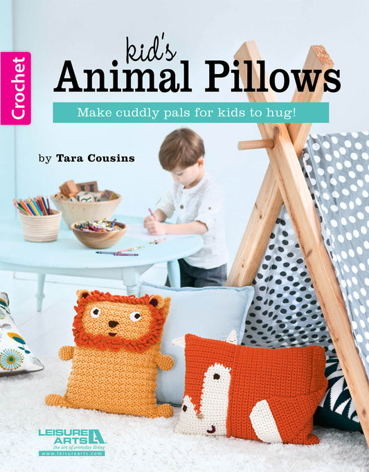 A child draws at a small table in a playroom with a teepee, polka dot blanket, and two crocheted animal pillows—a lion and fox—from the Kids Animal Pillows collection by Leisure Arts. The book cover also reads Kids Animal Pillows.