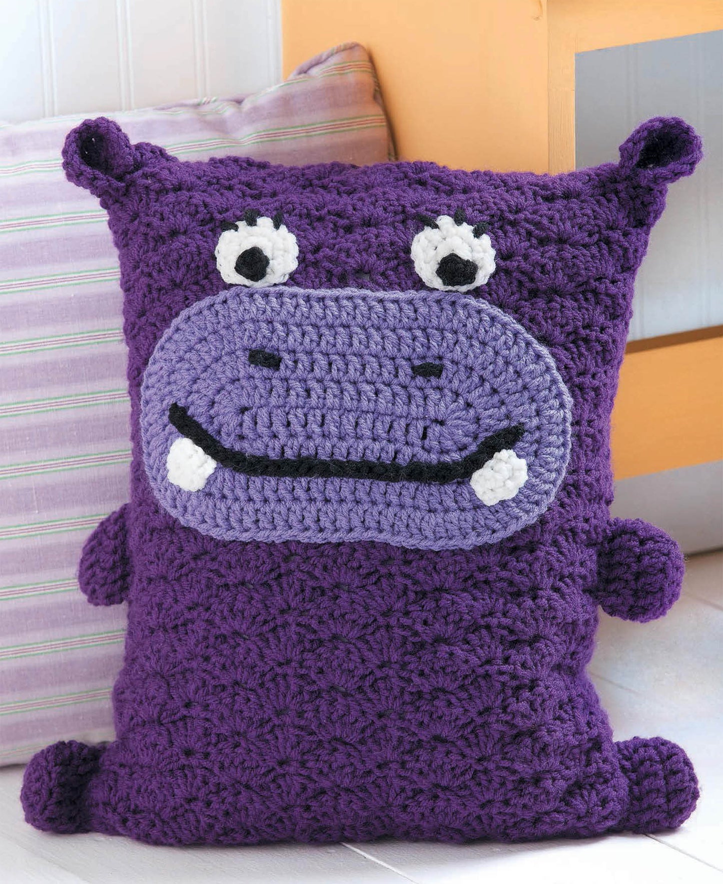 A purple rectangular Kids Animal Pillow by Leisure Arts, crocheted as a hippo with cute cartoon features and white teeth, leans against a striped cushion—perfect as an adorable decorative accessory for kids.