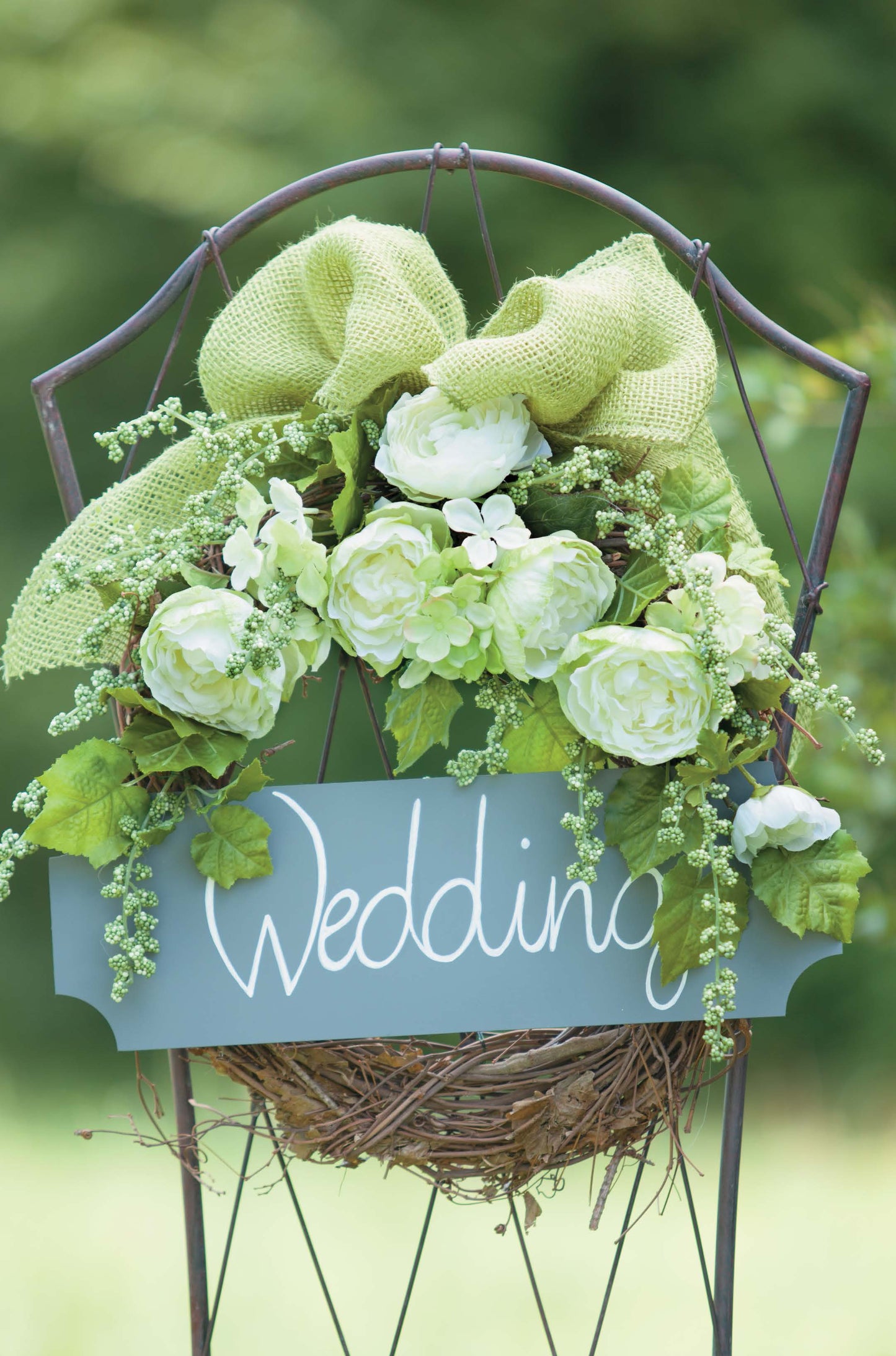The Rustic Wedding Florals wreath by Leisure Arts features white and green flowers, lush greenery, and a large green bow on a metal stand, with a “Wedding” sign—a charming country wedding decoration set amid blurred greenery.