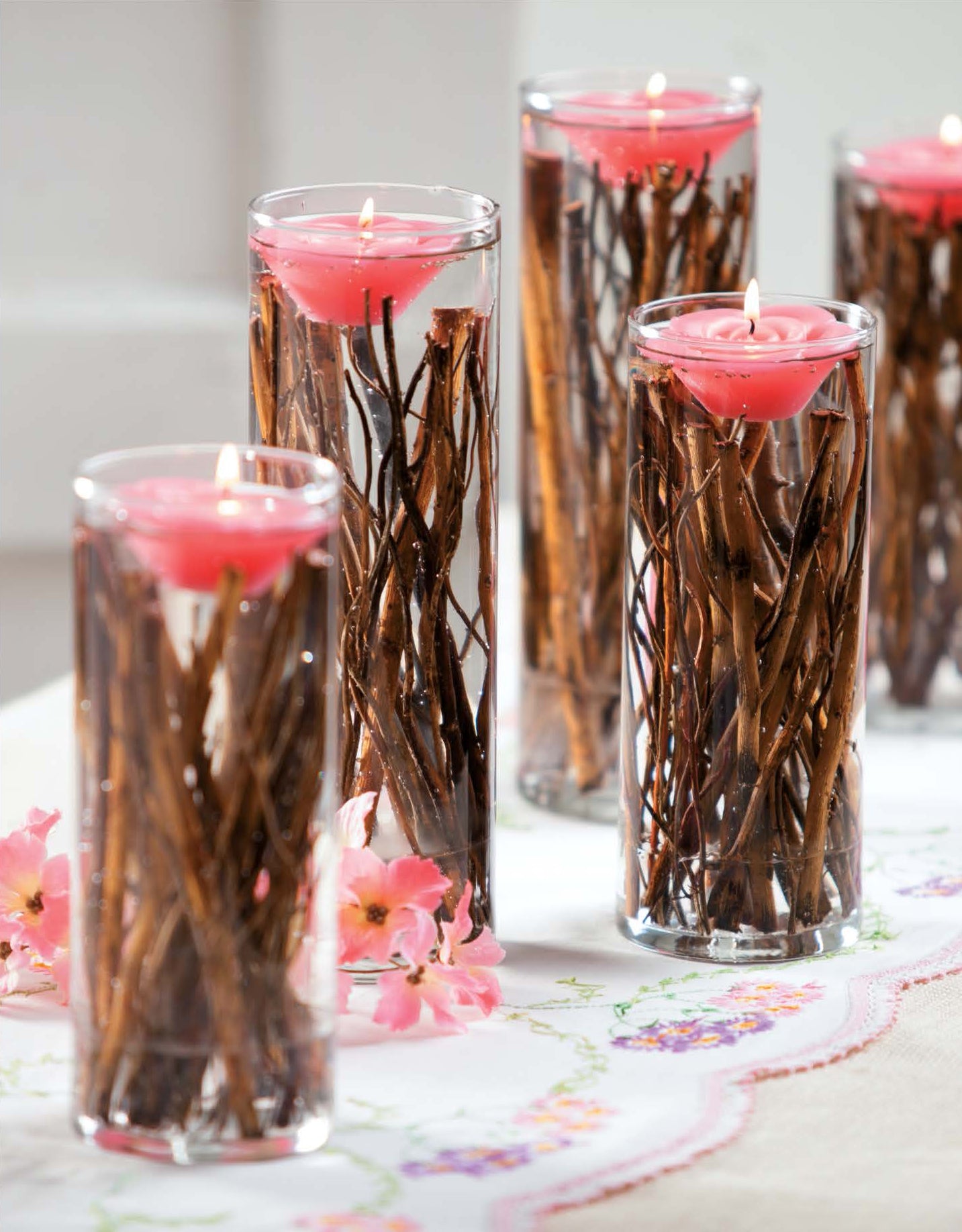 Leisure Arts' Rustic Wedding Florals features glass vases with water, twigs, and pink floating candles for charming DIY wedding decor. Pink flowers scattered on an embroidered tablecloth add to the rustic elegance.