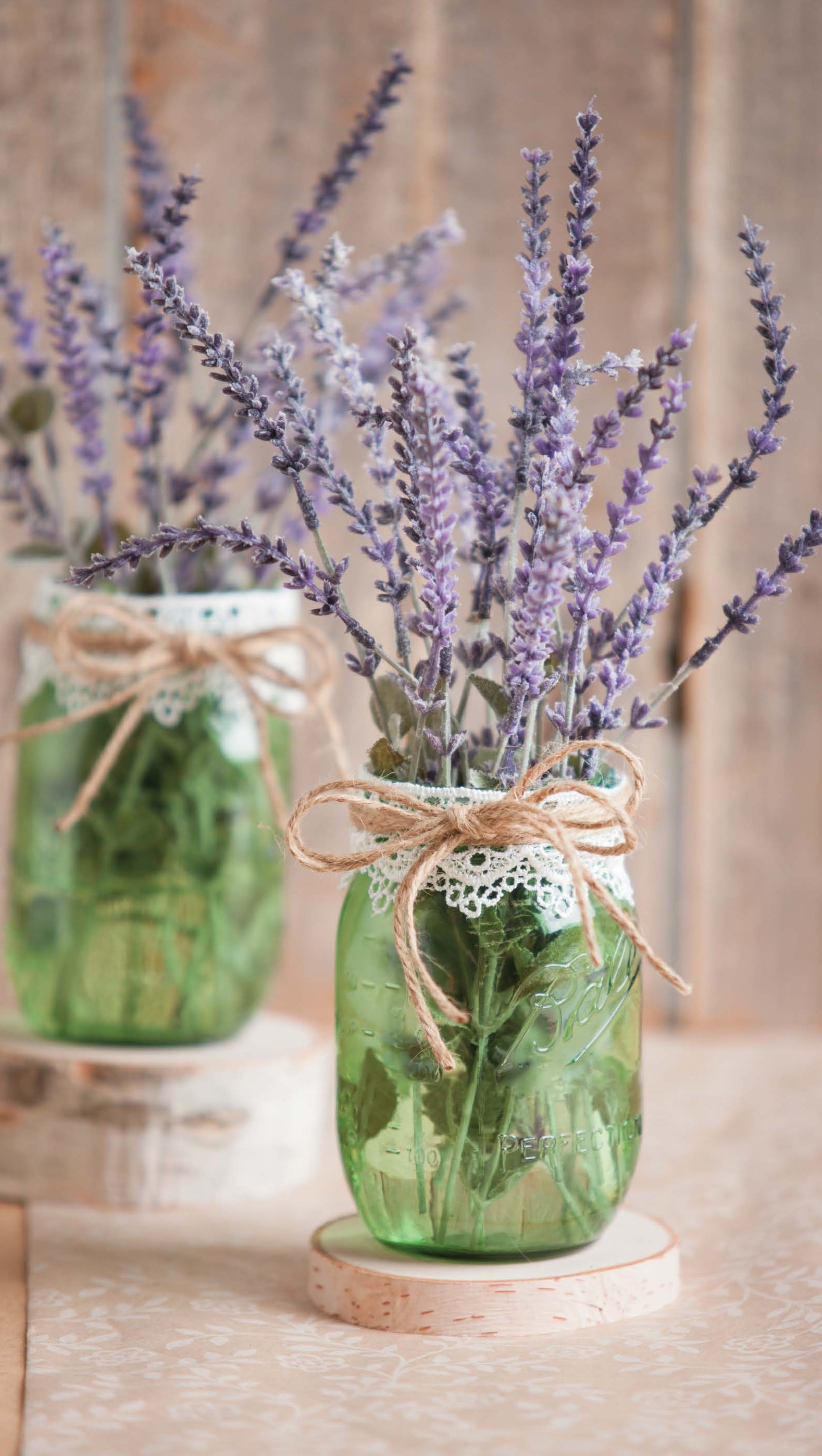 Rustic Wedding Florals by Leisure Arts features two green glass jars with lavender stems, lace and twine bows on wooden coasters—ideal DIY decor for country-themed or rustic weddings.