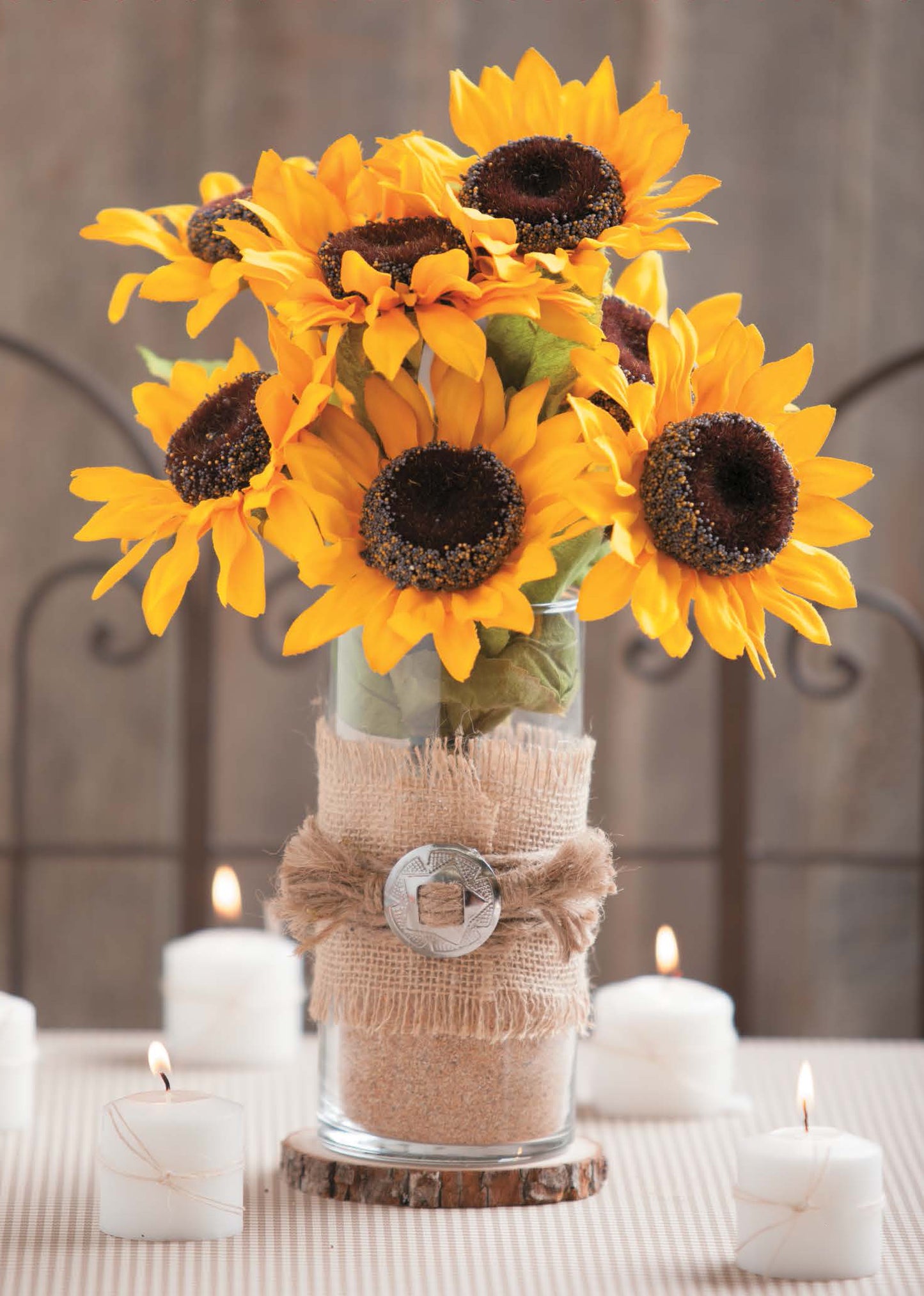 The Rustic Wedding Florals by Leisure Arts features a clear glass vase with vibrant sunflowers, burlap and a decorative button, surrounded by four lit white candles—ideal for rustic wedding decor. The background is softly blurred.