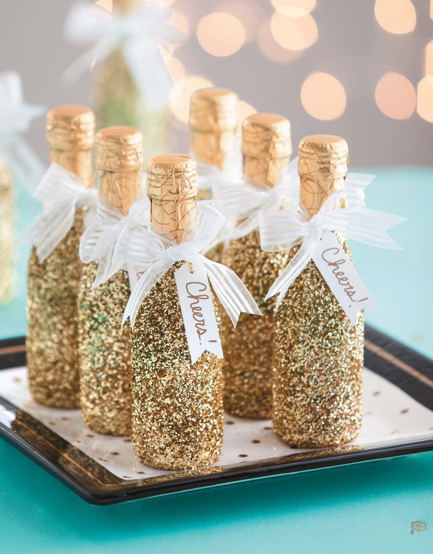 The Glam Wedding Favors Digital Download by Leisure Arts features small champagne bottles adorned with gold glitter, white bows, and Cheers! tags on a black tray—charming, budget-friendly DIY gifts in a warm bokeh-lit setting.
