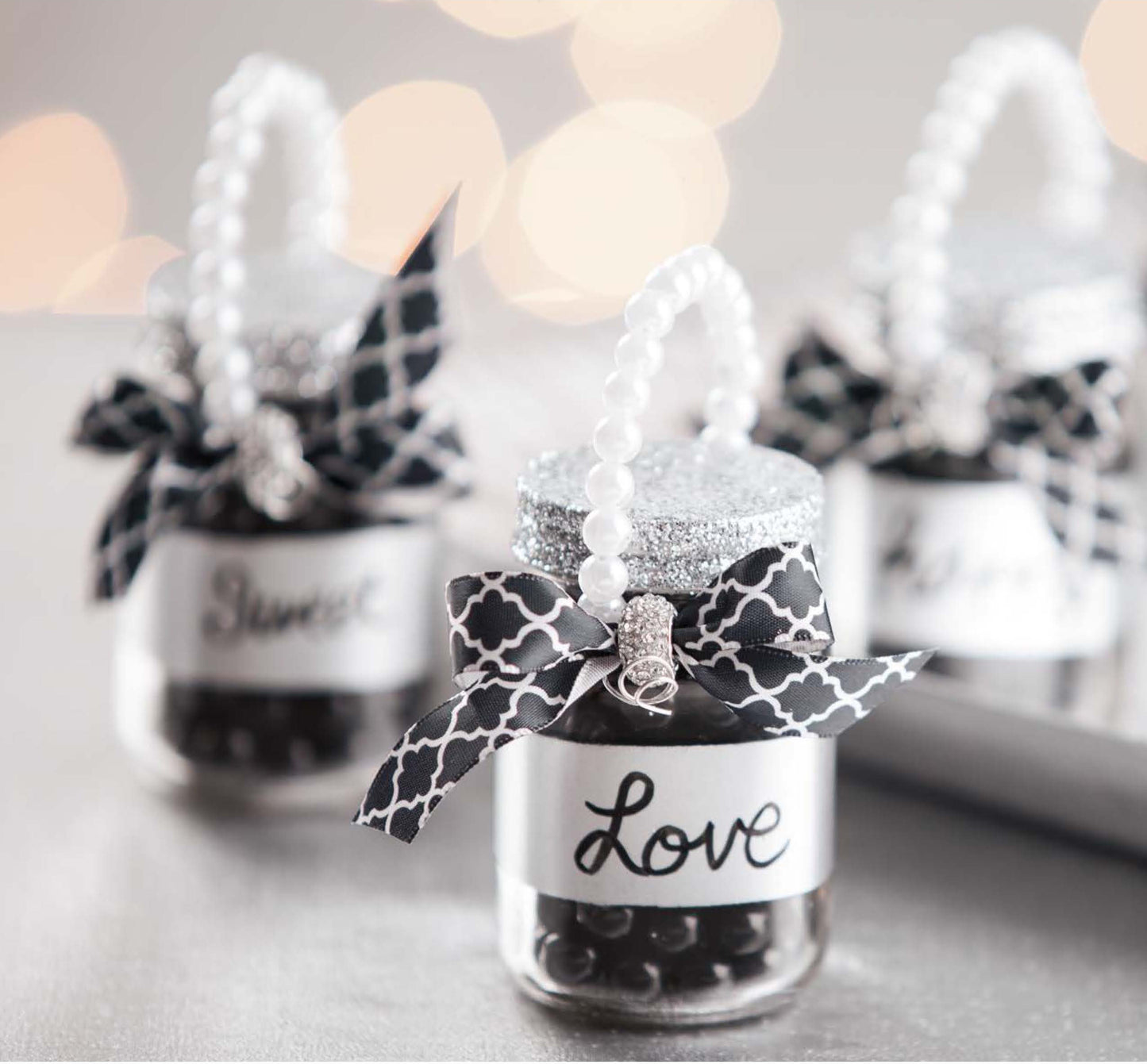 The Glam Wedding Favors Digital Download by Leisure Arts features three small jars with black and white bows, glitter lids, pearl handles, and “Love” labels. Fill with candies for affordable DIY gifts or favors for any celebration.