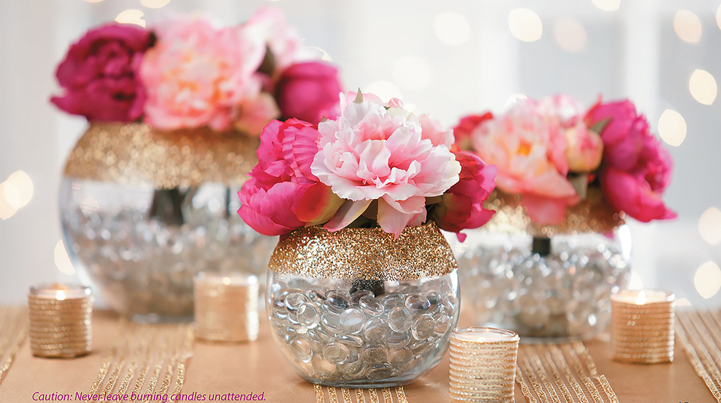 The "Glam Wedding Florals" kit by Leisure Arts features round glass vases with gold glitter tops and pink-white flowers, accented by tealight candles on a gold tablecloth—perfect for dazzling DIY wedding decor.