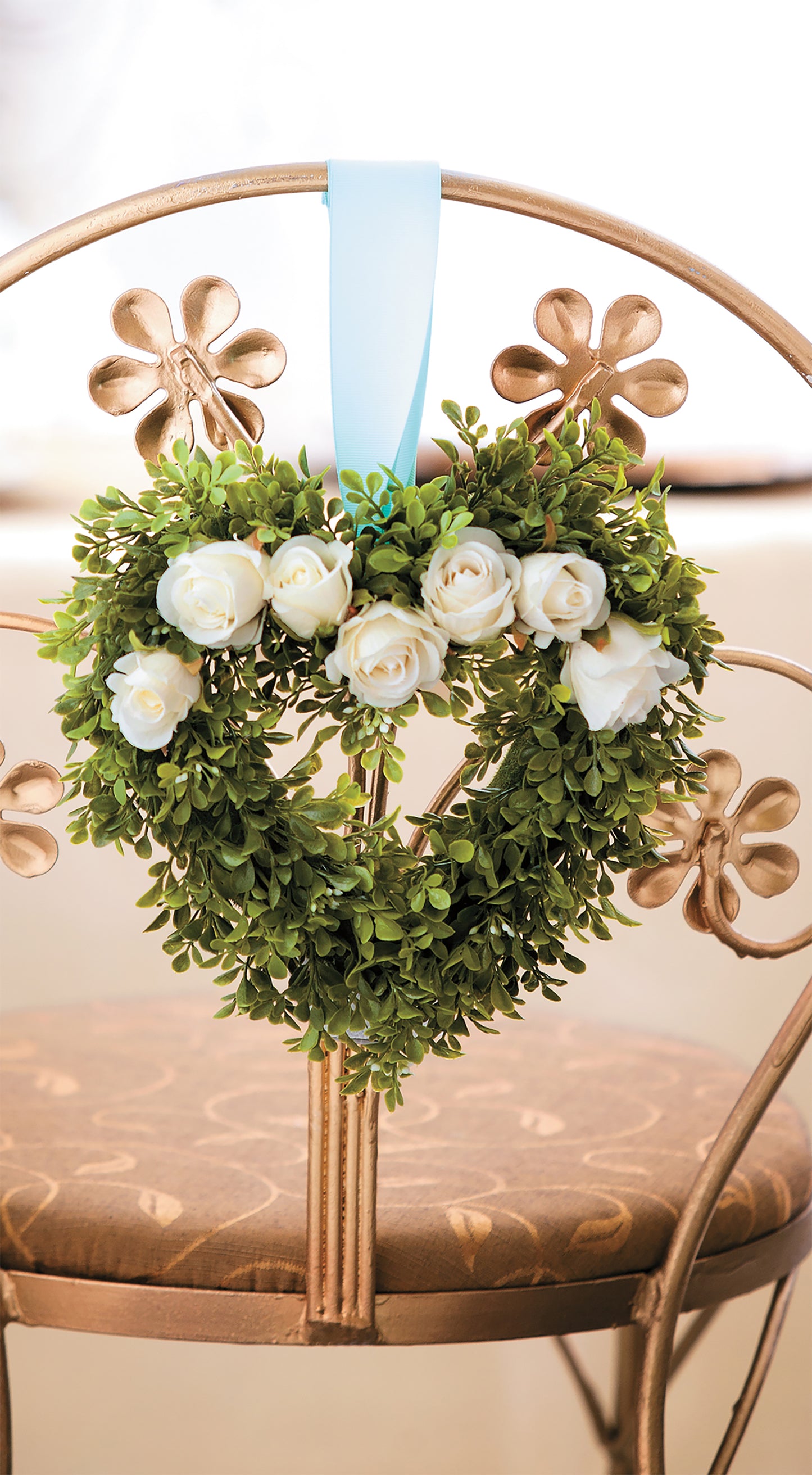 The Glam Wedding Florals by Leisure Arts features a heart-shaped green leaf and white rose wreath, perfect for DIY wedding decor, hanging from a light blue ribbon on a decorative metal chair with a floral motif.