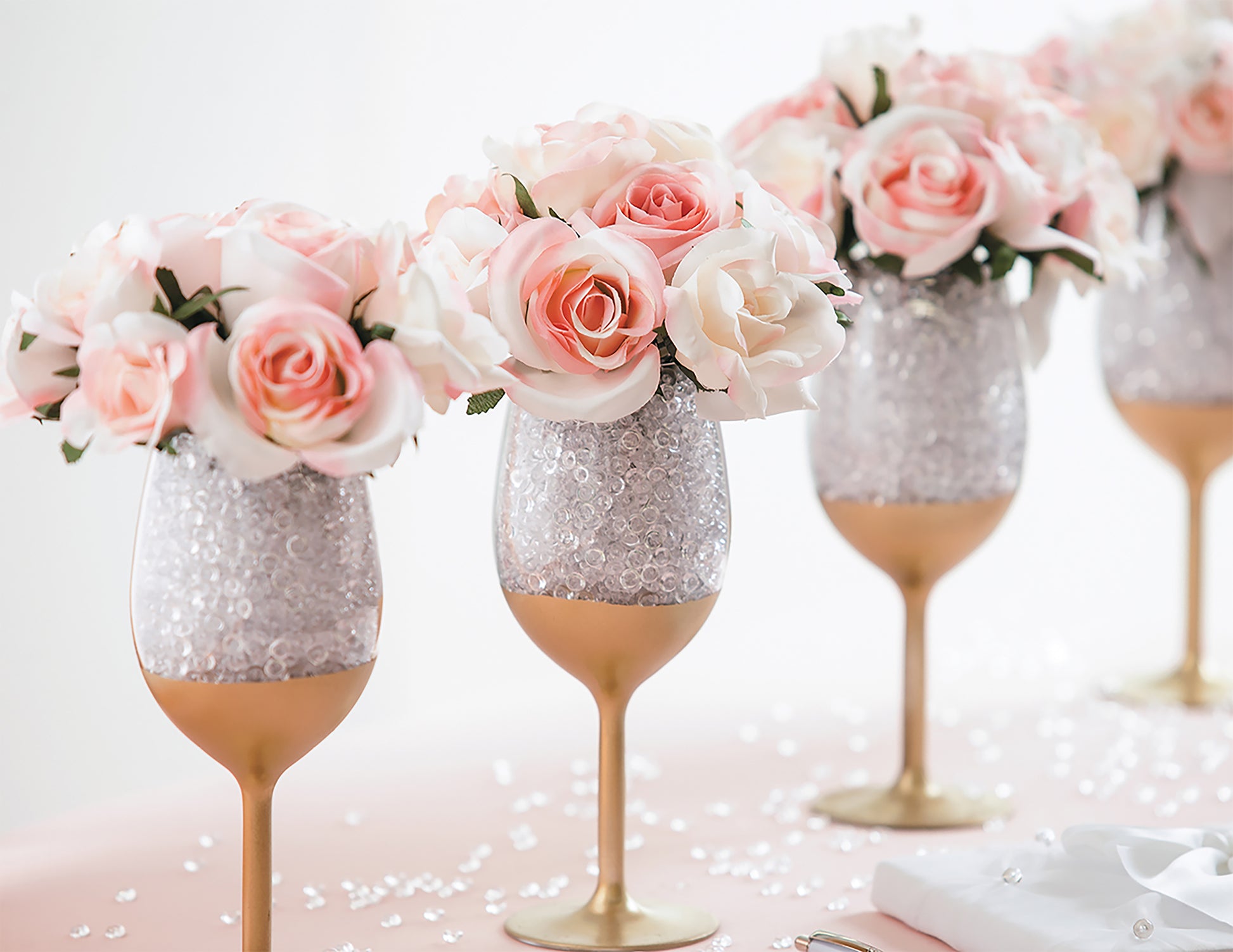 Create an elegant centerpiece with Glam Wedding Florals by Leisure Arts—four gold-stemmed glasses with clear beads and pink and white faux roses on a blush tablecloth, ideal for DIY wedding decor or beginner floral arrangements.