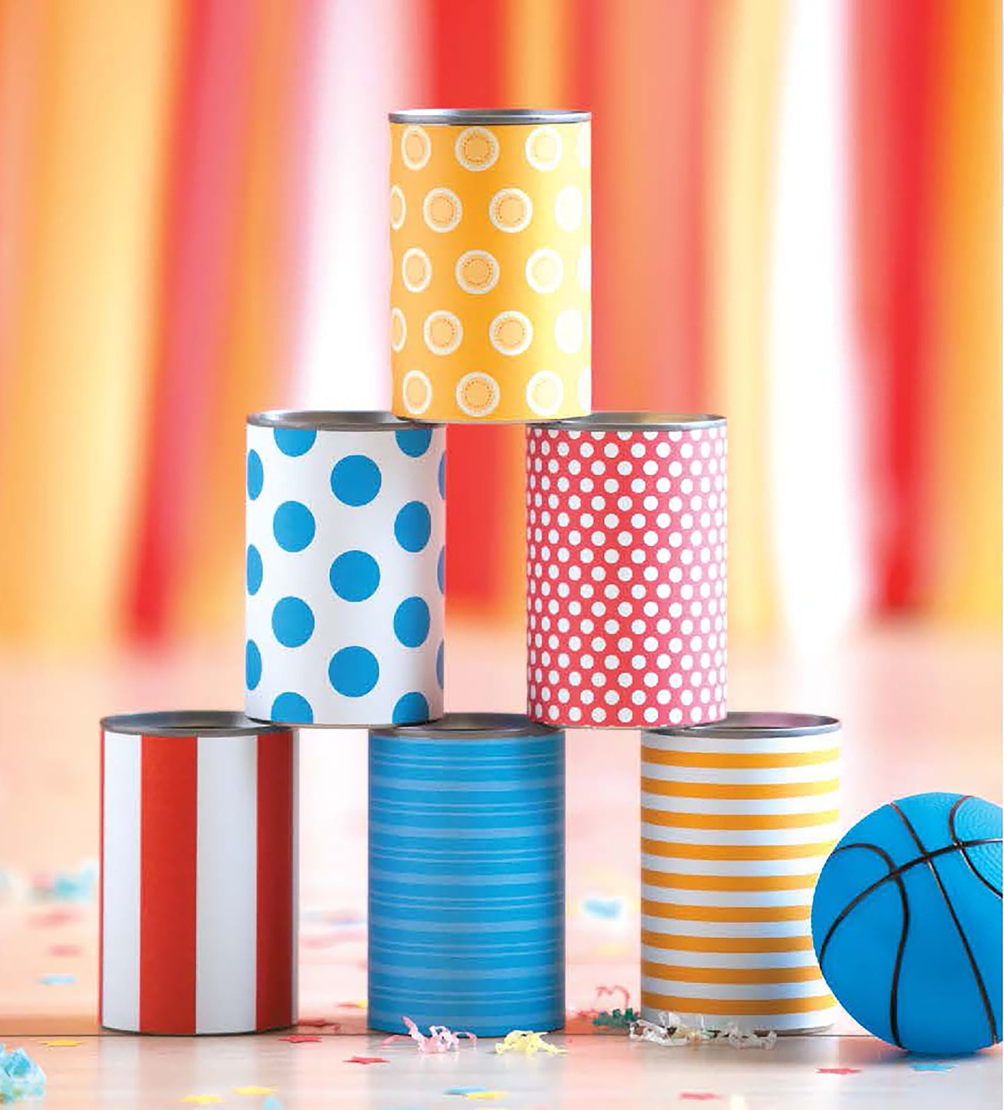 Six colorful Celebrate Birthdays cans from Leisure Arts, with vibrant patterns, are stacked pyramid-style amid confetti and a blue basketball—ideal DIY party crafts for kids or adults—set against warm striped backgrounds.