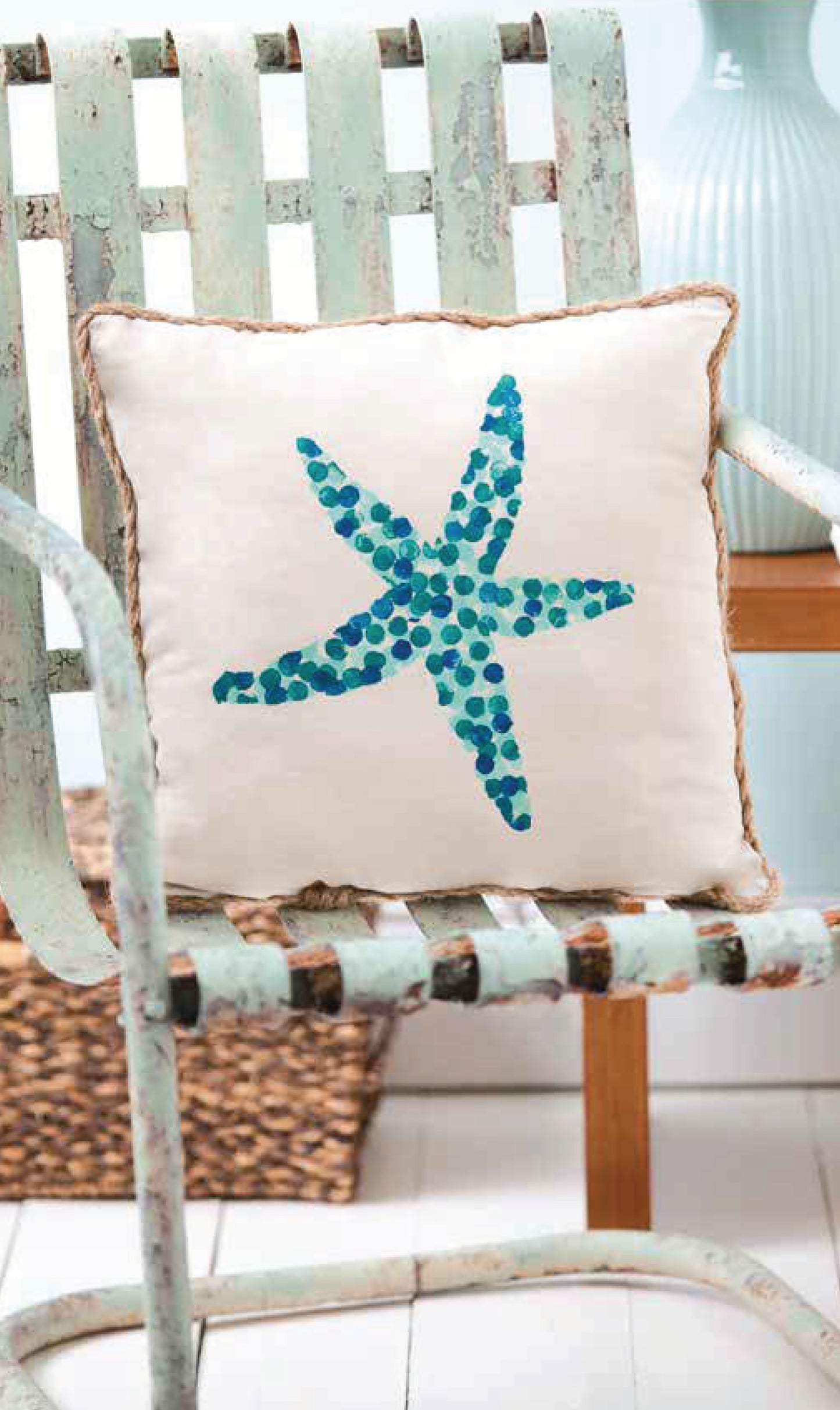 The "Painting Basics" book by Leisure Arts rests on a distressed pale green metal chair. Paired with a wooden table and light blue vase, it inspires creative DIY projects like painted rocks.