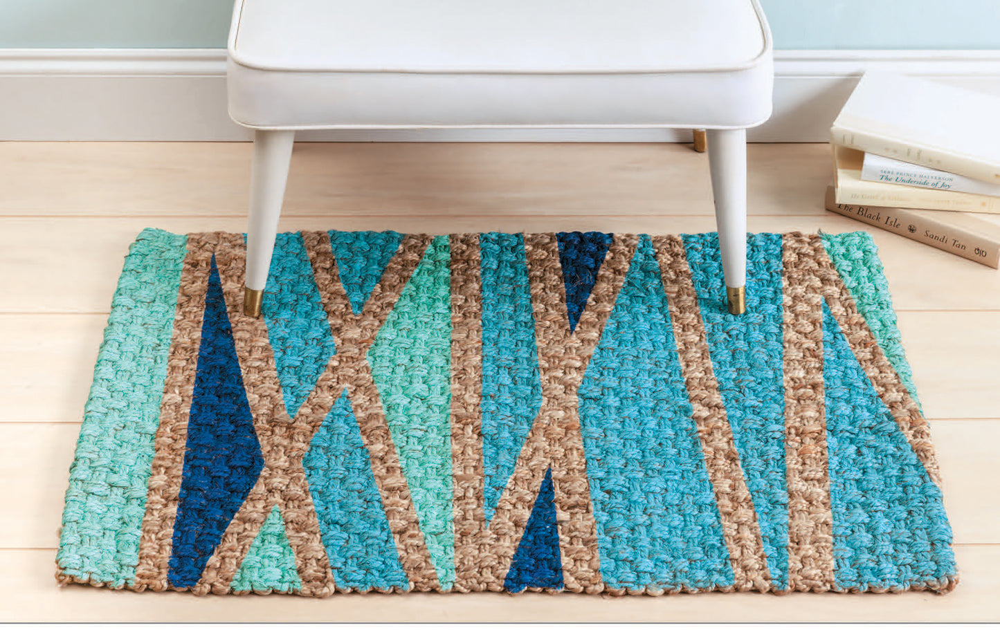 A white bench sits on a small multicolored geometric rug atop light wood floors. Stacked books and the "Stenciling Basics Digital Download" by Leisure Arts nearby spark ideas for creative home decor.