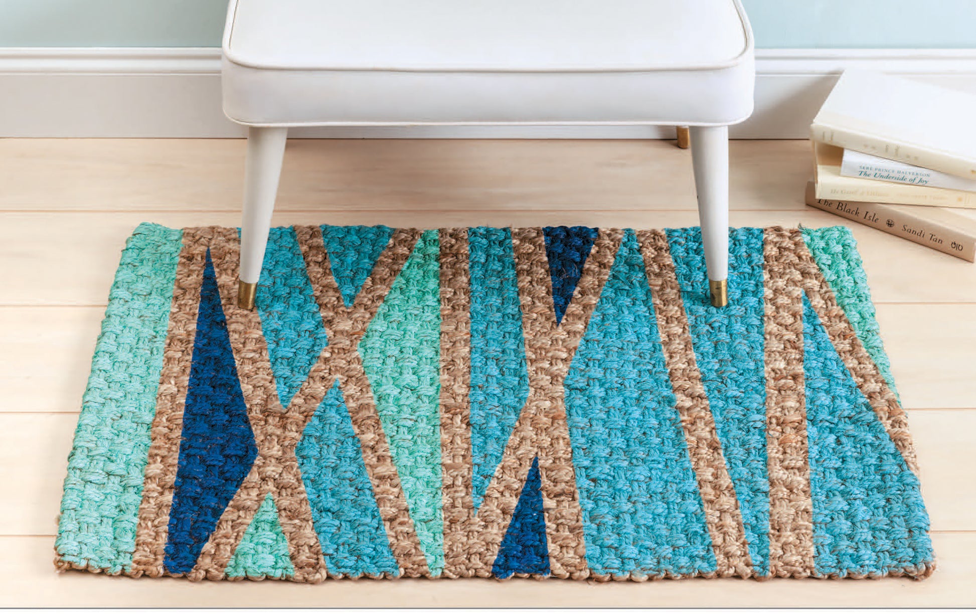 A white bench sits on a small multicolored geometric rug atop light wood floors. Stacked books and the "Stenciling Basics Digital Download" by Leisure Arts nearby spark ideas for creative home decor.