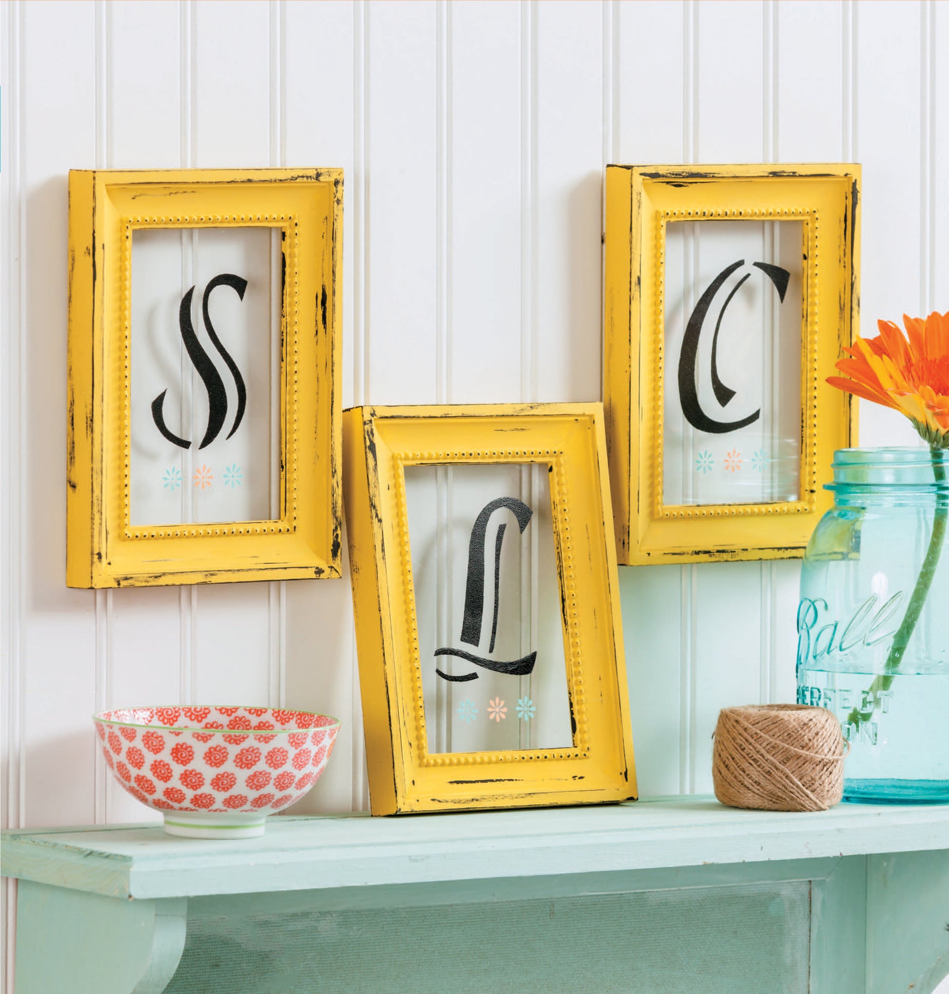 Stenciling Basics Digital Download by Leisure Arts lets you create monogrammed decor, like three distressed yellow frames on a mint shelf with floral accents and more against white beadboard for a personalized home touch.