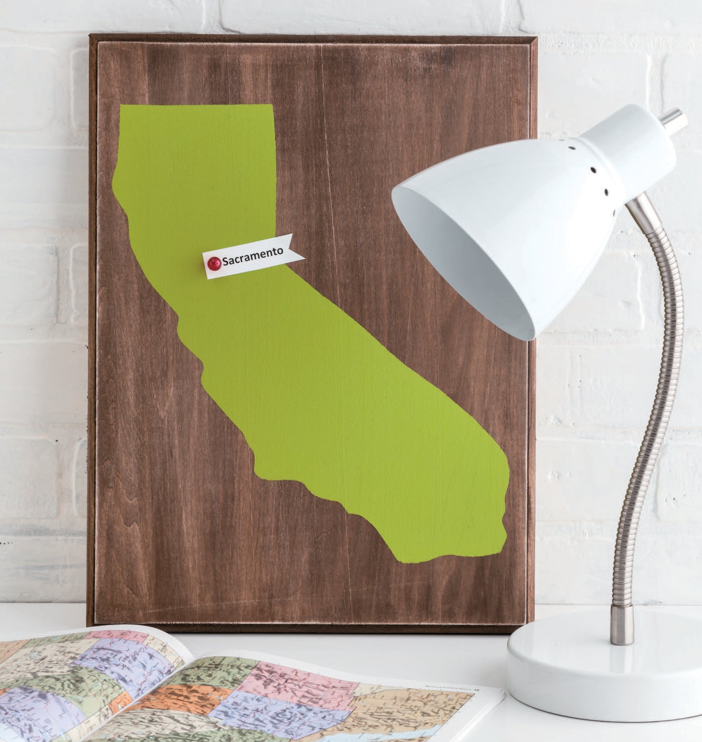 The Stenciling Basics Digital Download by Leisure Arts inspires creative projects—a wooden plaque with a green California silhouette and Sacramento pin adds thoughtful decor beside an atlas and desk lamp.