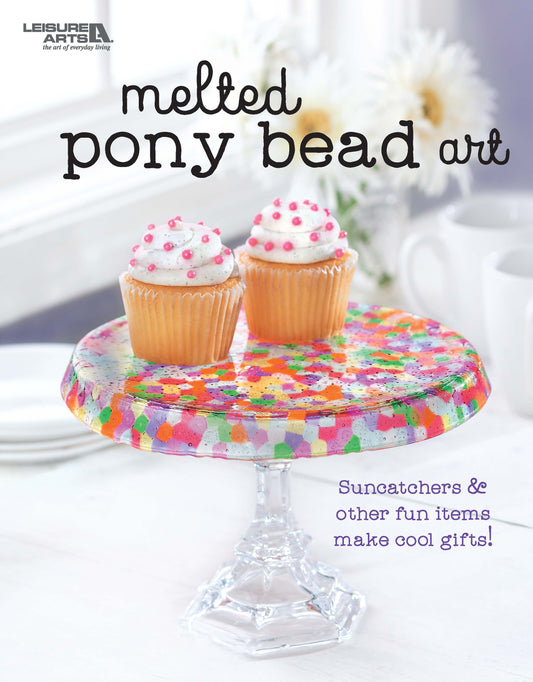 Two cupcakes with white frosting and pink sprinkles sit on a colorful Melted Pony Bead Art cake stand by Leisure Arts, set against a light background—perfect for displaying beginner crafts or creative melted bead gift ideas.