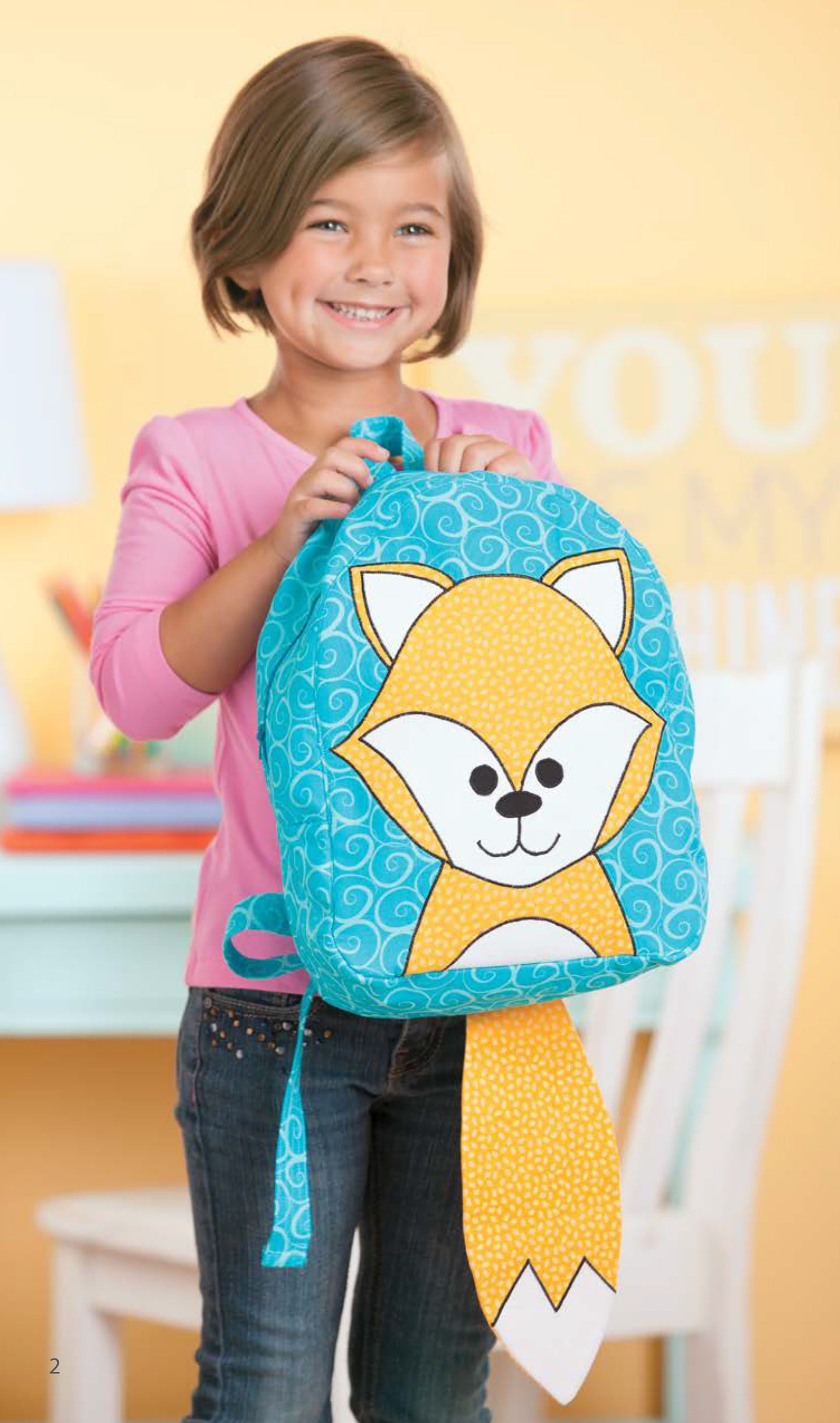 A smiling young girl with short brown hair holds a Leisure Arts Woodland Creatures Backpack featuring a cute yellow fox, standing in a colorful room filled with playful applique patterns.
