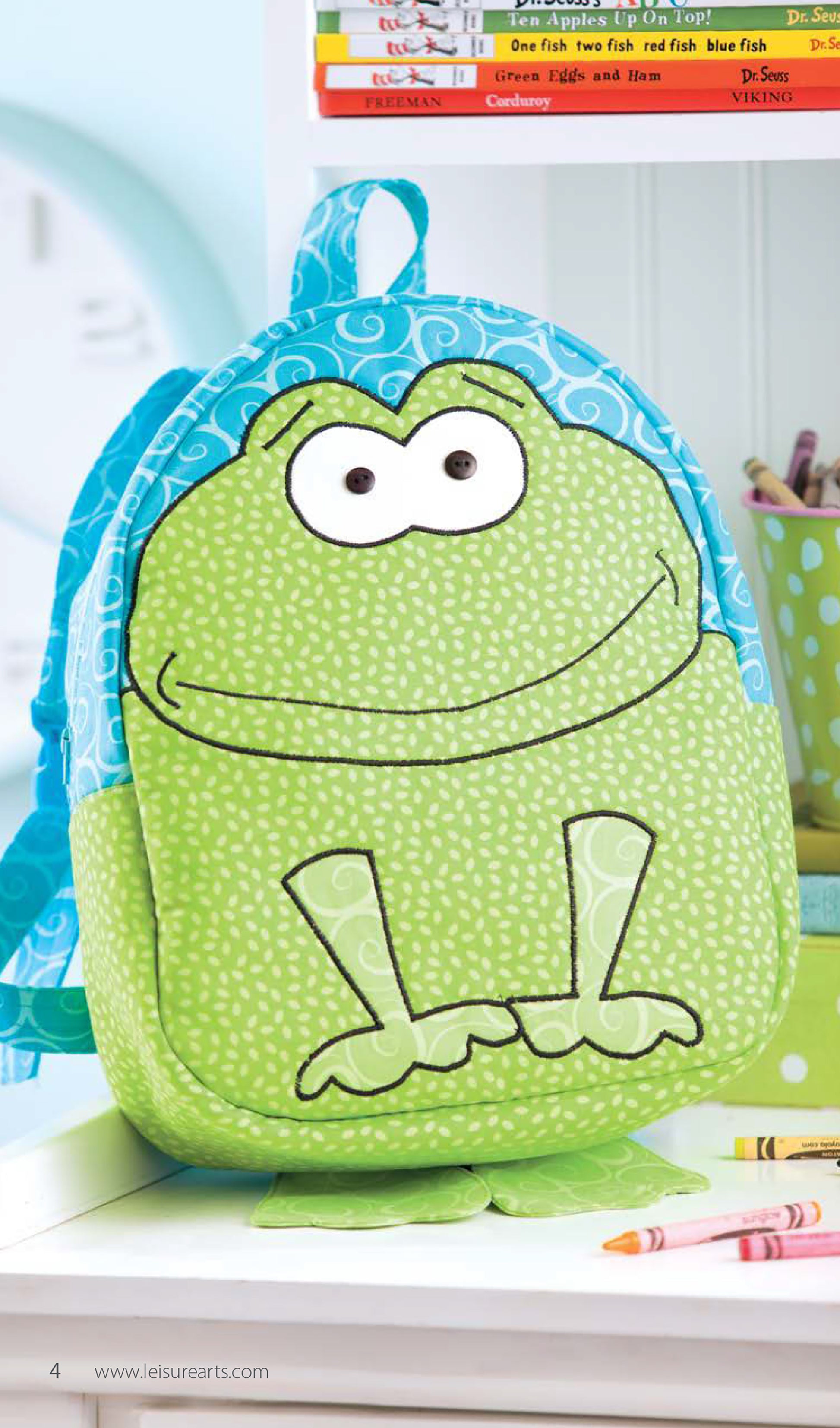 The Leisure Arts Woodland Creatures Backpacks kids' carryall features a cute green frog design. Shown on a white surface with crayons, books, and a green polka dot cup of pencils in the background.