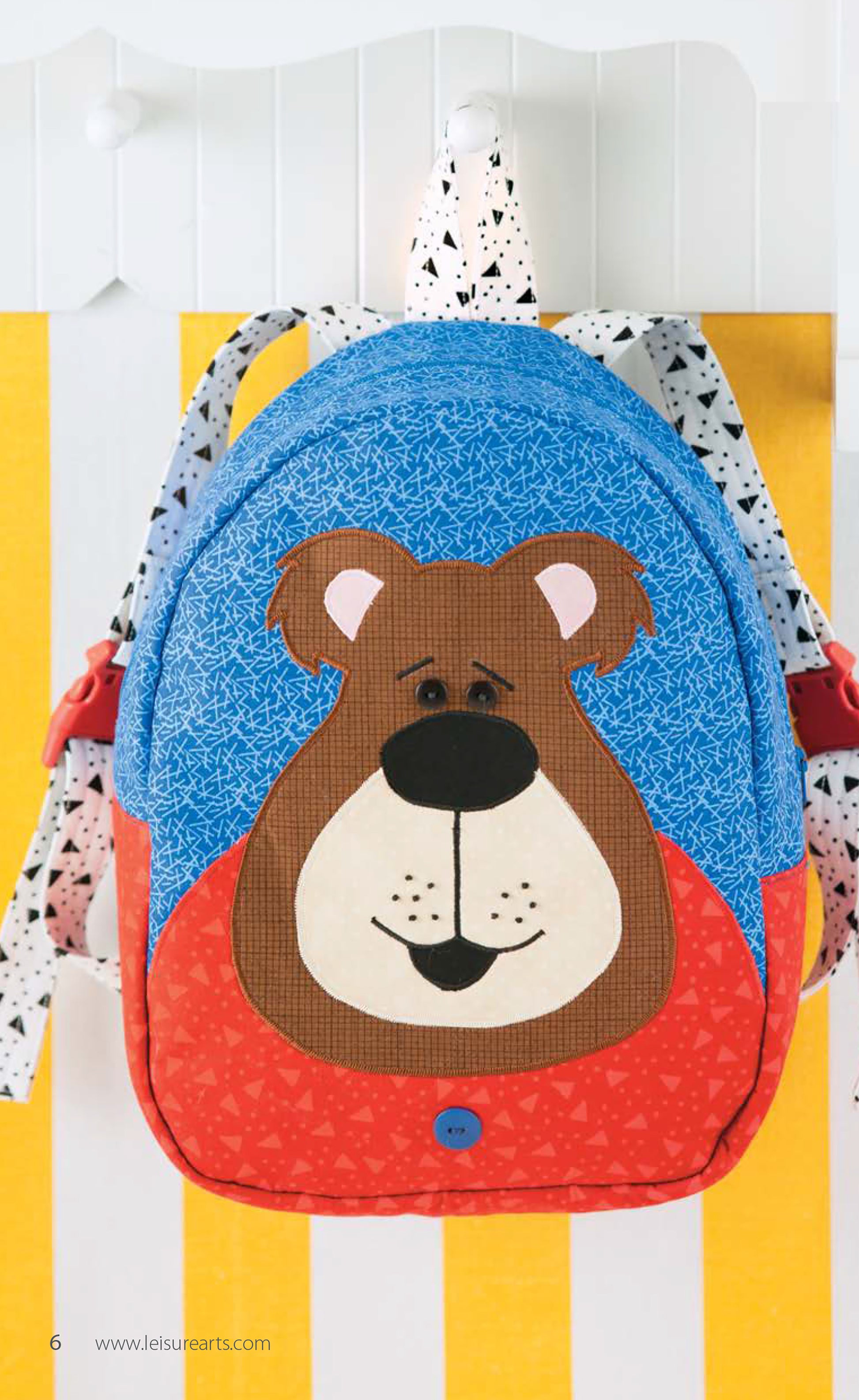 The Woodland Creatures Backpacks by Leisure Arts, featuring a playful bear face in blue, red, and brown applique, hangs on a white hook against a yellow and white striped background.