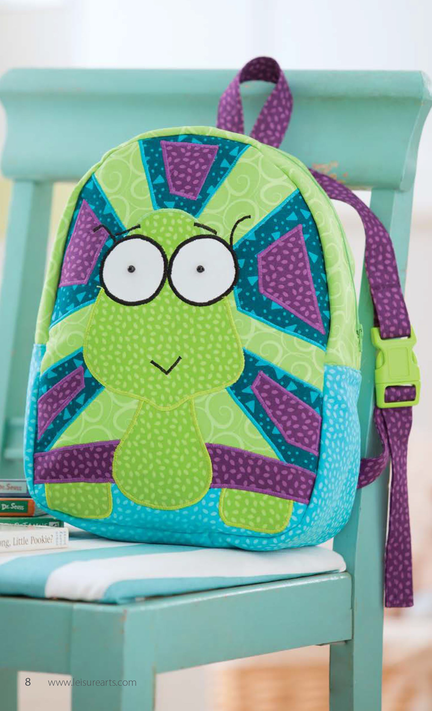 The Leisure Arts Woodland Creatures Backpacks, designed for kids with a cartoon frog face and colorful green, blue, and purple appliques, sits on a light turquoise chair beside a stack of books.