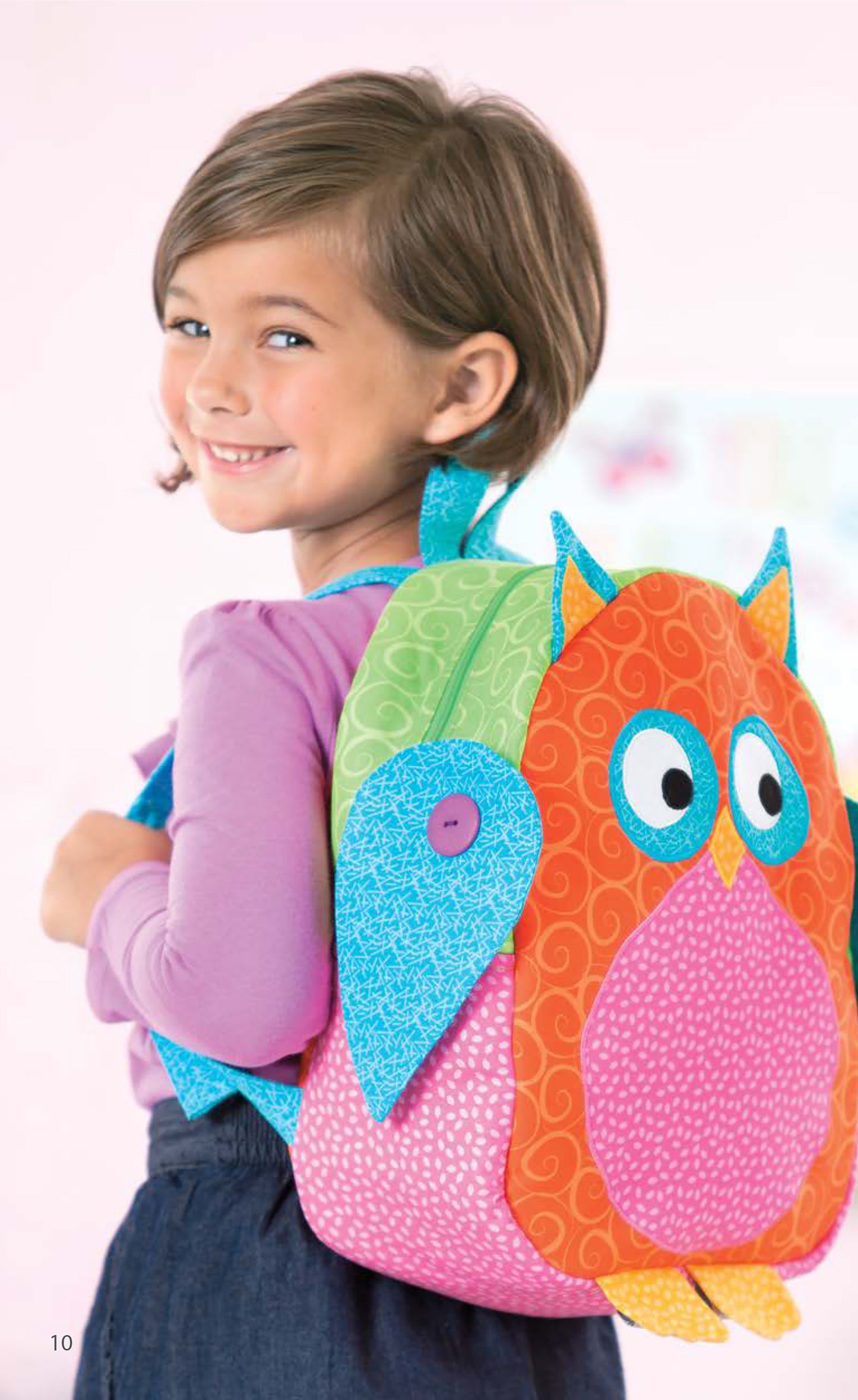 A young child with short brown hair smiles while wearing a Leisure Arts Woodland Creatures Backpack, featuring vibrant orange, pink, blue, and green fabric with large button eyes and playful owl applique patterns.