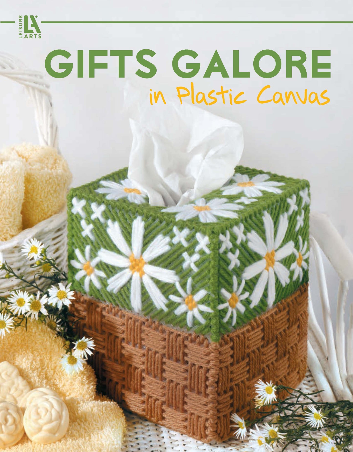 A tissue box with a handmade plastic canvas cover featuring white daisies and green leaves sits beside yellow towels and daisies. "Gifts Galore in Plastic Canvas Digital Download" by Leisure Arts is perfect for handmade gift ideas.