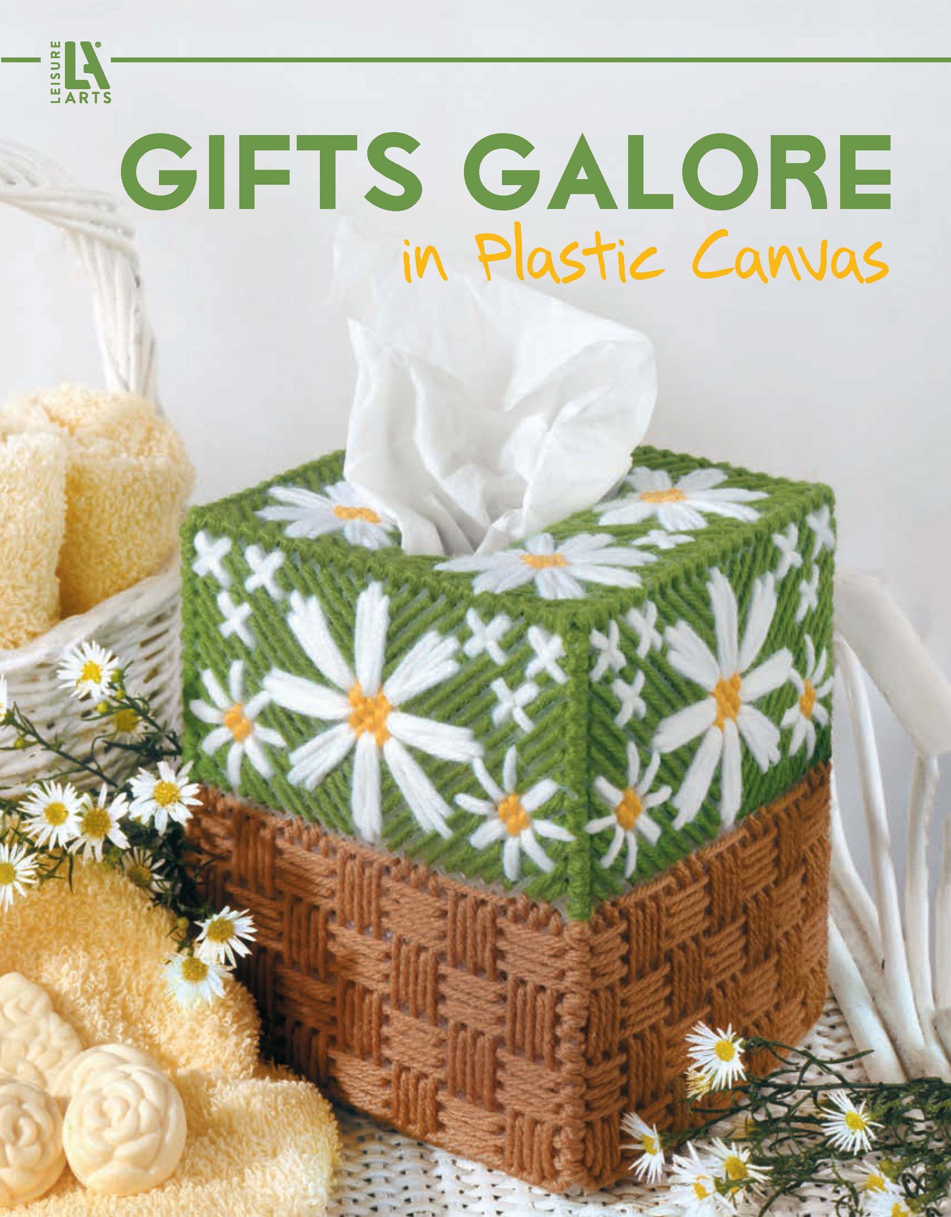 A tissue box with a handmade plastic canvas cover featuring white daisies and green leaves sits beside yellow towels and daisies. "Gifts Galore in Plastic Canvas Digital Download" by Leisure Arts is perfect for handmade gift ideas.