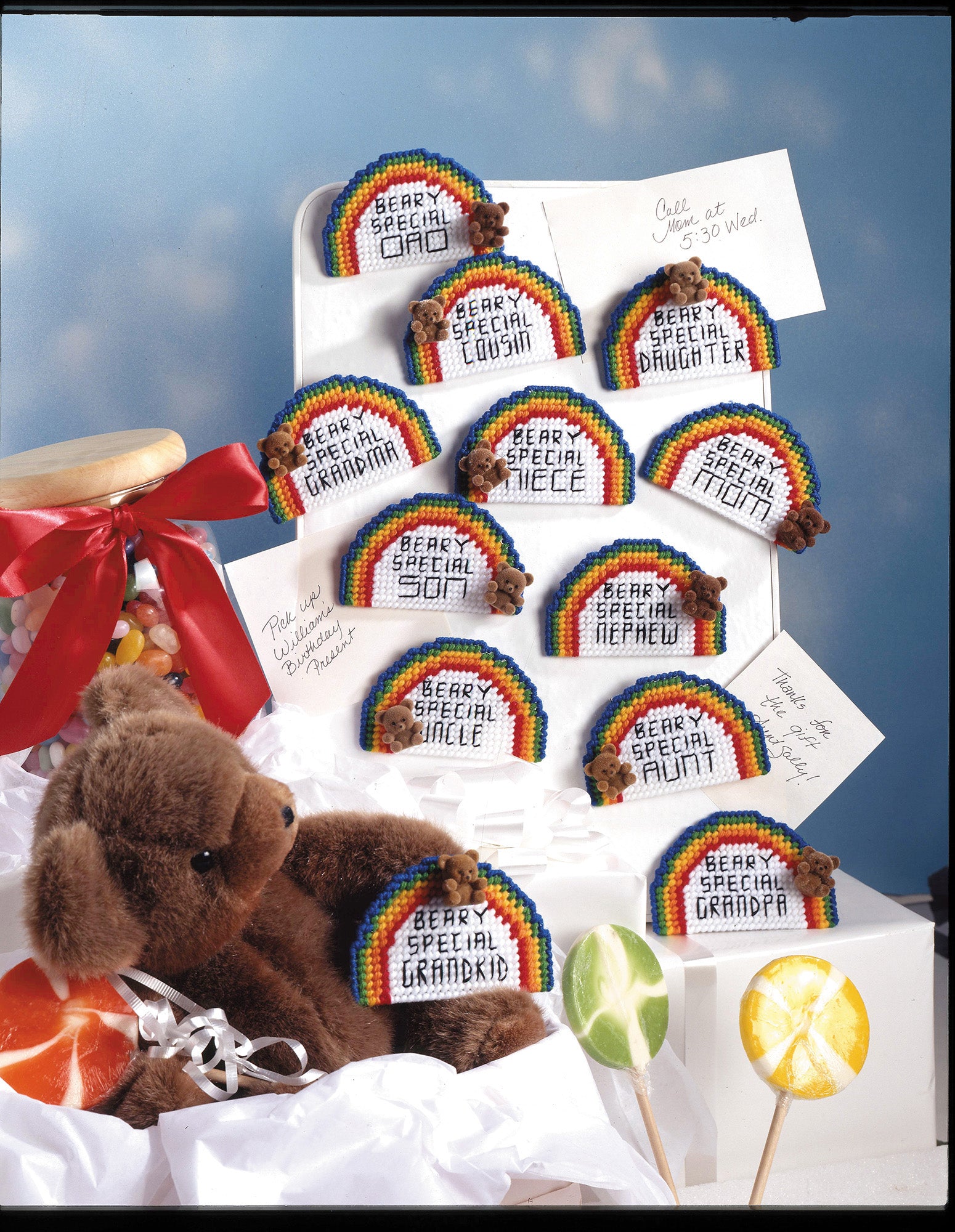 Gifts Galore in Plastic Canvas Digital Download by Leisure Arts features rainbow plaques with messages like Beary Special Mama and Beary Special Grandpa, plus teddy bears, gifts, and lollipops on a blue sky background.