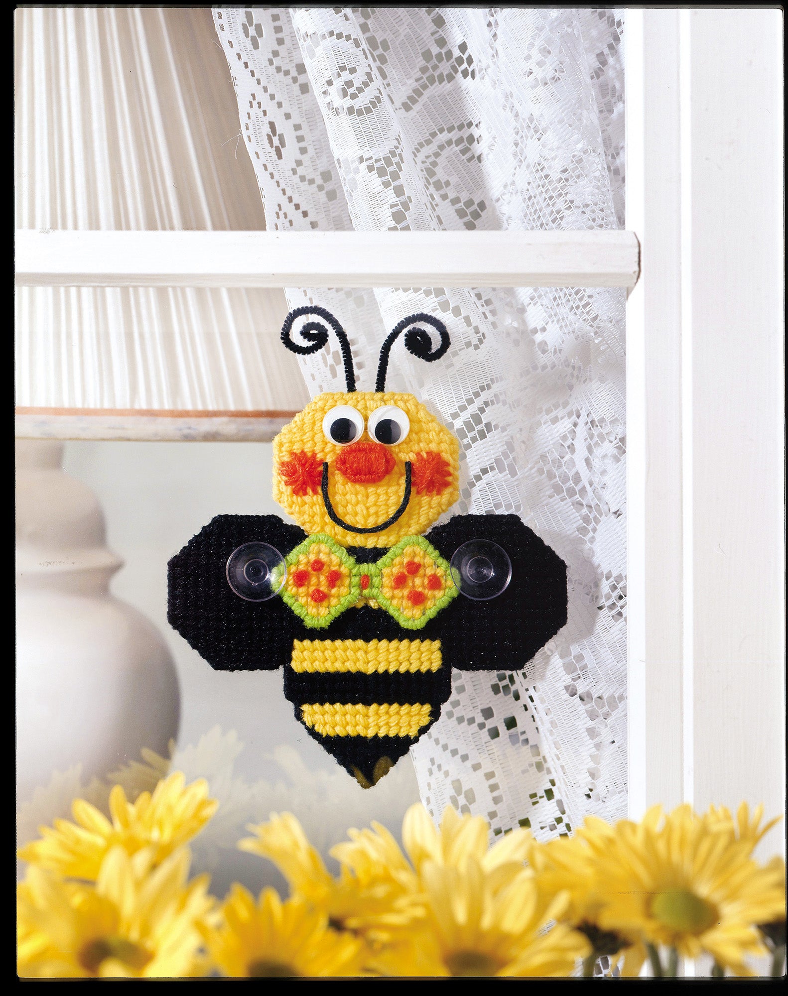 A cheerful, handmade bee from the Leisure Arts "Gifts Galore in Plastic Canvas" digital download brightens the window, surrounded by yellow flowers and lace curtains—perfect for easy crafts or handmade gifts.