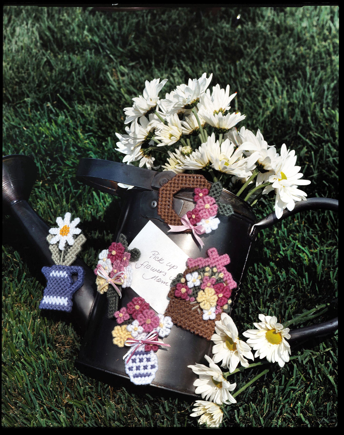 A black watering can with white daisies sits on grass, adorned with colorful floral and basket motifs from Gifts Galore in Plastic Canvas by Leisure Arts—perfect for quick crafts or handmade gift ideas. Digital download available.
