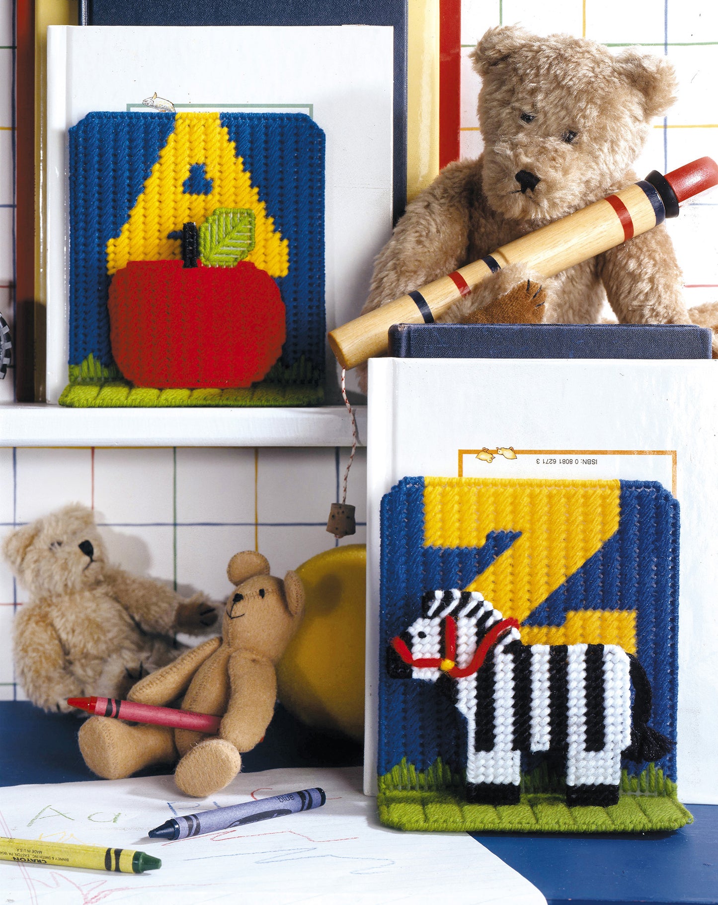 Leisure Arts’ Gifts Galore in Plastic Canvas Digital Download shows three teddy bears with themed crafts—apple and A, zebra and Z. Surrounded by crayons, drawings, a recorder, and a yellow ball near a grid wall, it's quick and easy fun!.