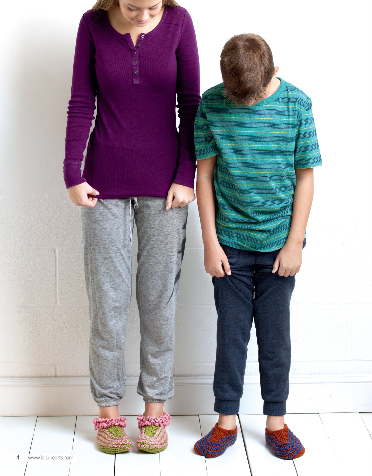 A woman and a boy stand indoors in casual clothes, admiring their feet in cozy slippers made from the "Slipper for the Family Digital Download" by Leisure Arts—an ideal beginner crochet project for everyone.