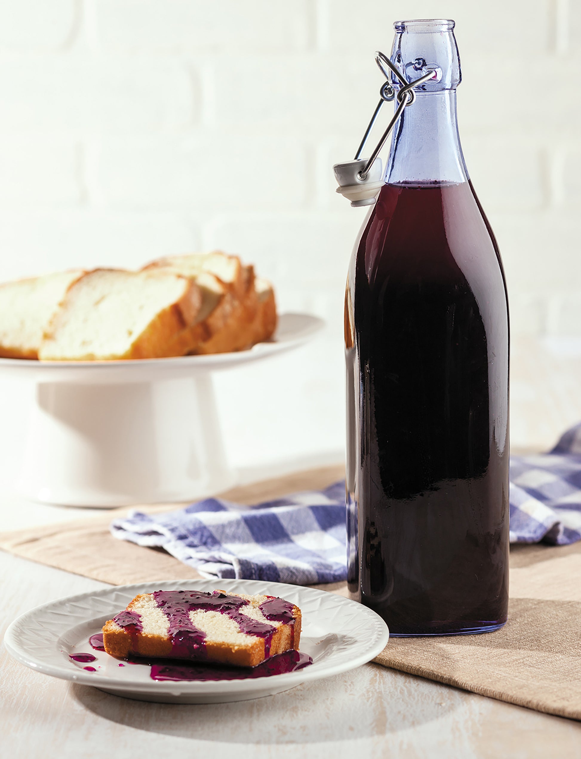 A copy of Leisure Arts' "Canning Made Easy" sits beside a plate with a slice of pound cake topped with purple jam; behind it, more cake rests on a white stand next to a blue checked cloth.