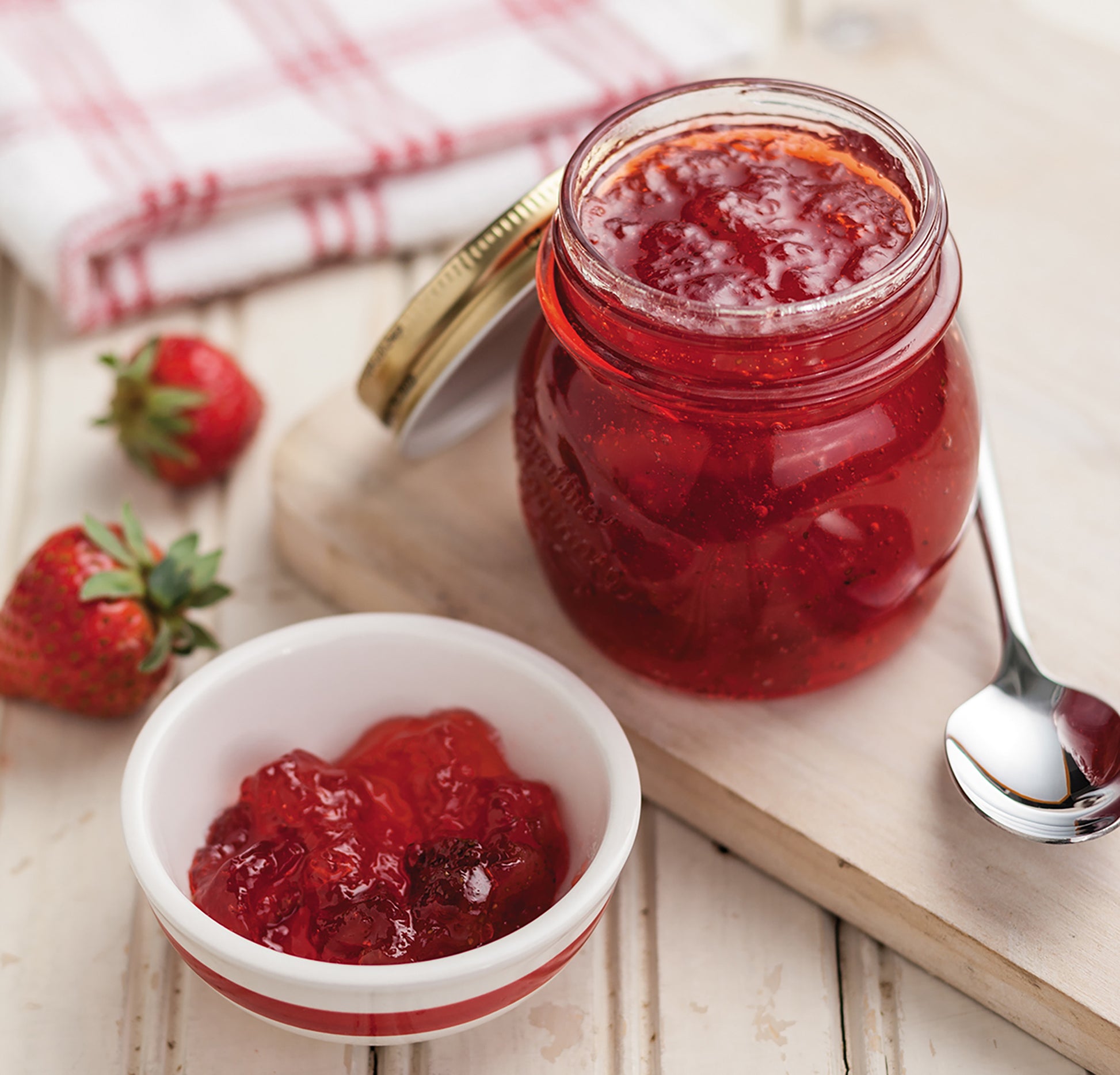 A glass jar of strawberry jam with the lid off sits on a wooden board beside a spoon, fresh strawberries, and a red-and-white checkered cloth—perfect for homemade jams or pickles inspired by Canning Made Easy from Leisure Arts.