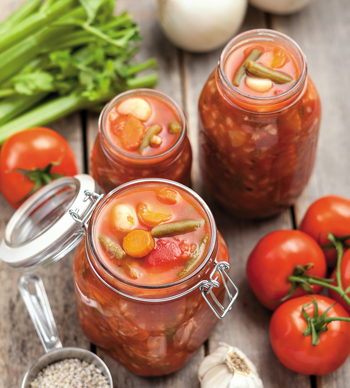 Three jars of homemade vegetable soup from Leisure Arts' Canning Made Easy are displayed on a wooden table with fresh tomatoes, celery, garlic, onions, grains, and pickles.