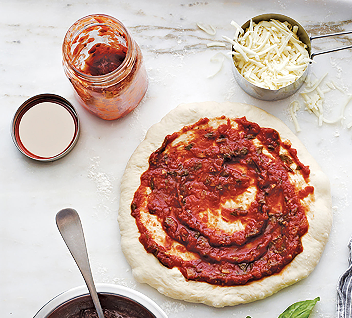 A pizza dough with tomato sauce rests on a floured surface beside an open jar of Canning Made Easy by Leisure Arts salsa, a metal cup of shredded cheese, and a spoon.