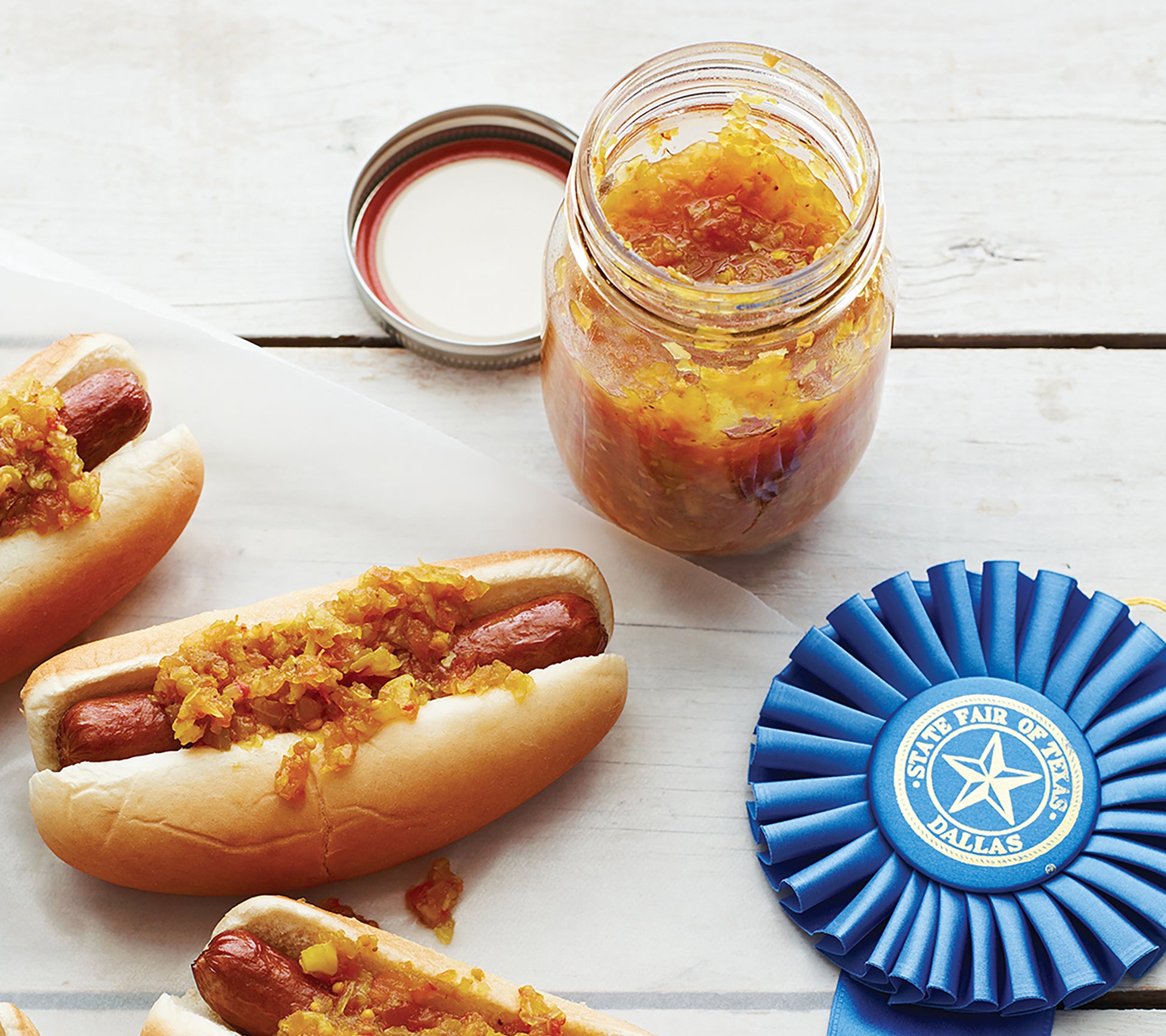 Hot dogs in buns topped with chunky relish and pickles, next to an open jar of homemade relish made using Canning Made Easy by Leisure Arts, and a blue first-place ribbon from the State Fair of Texas, Dallas.