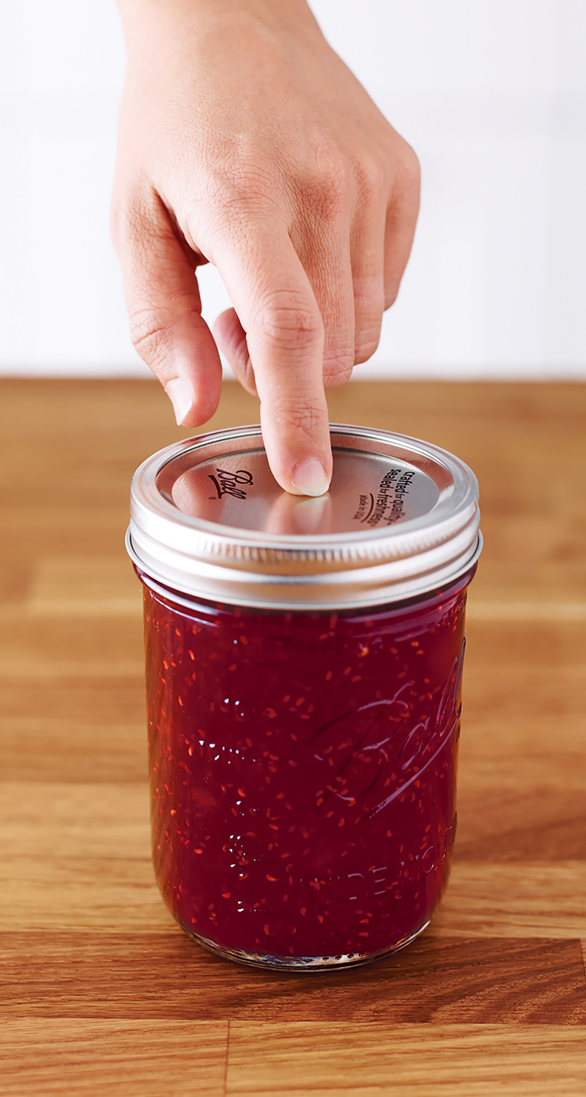 A hand presses down on the metal lid of a glass jar filled with red raspberry jam, using Canning Made Easy by Leisure Arts, on a wooden surface with other homemade jams.