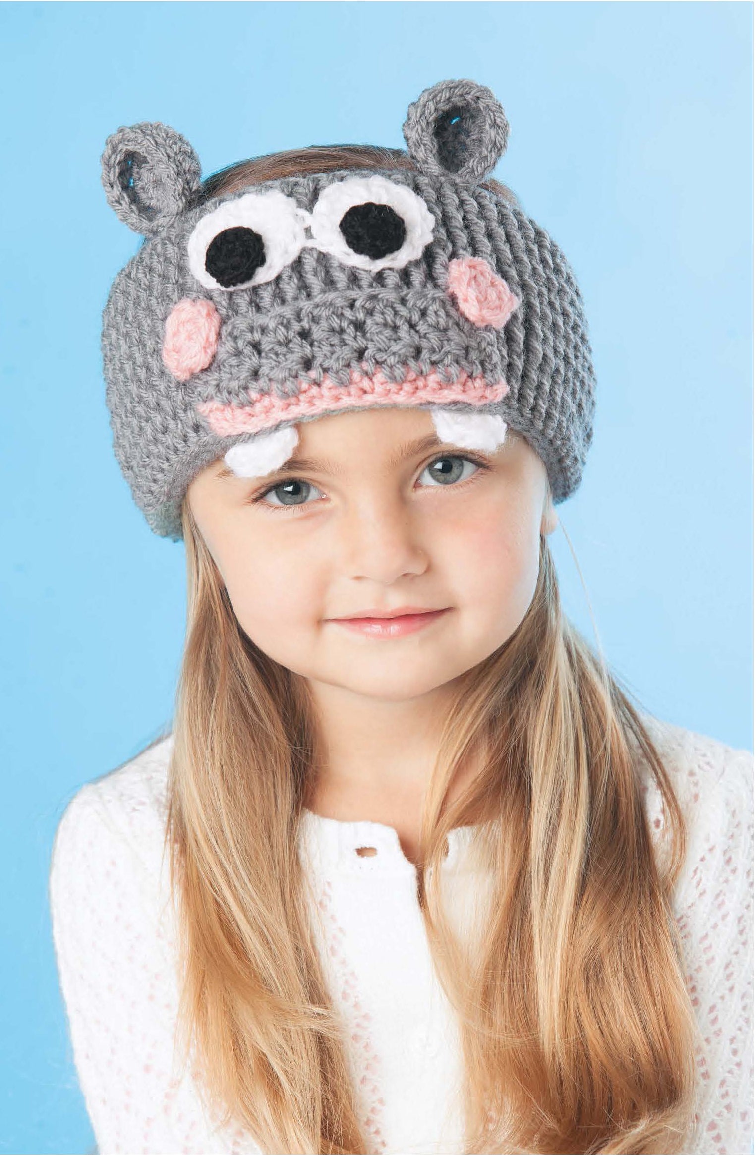 A young girl with long blonde hair wears the Leisure Arts Little Animals Headgear—a crocheted gray hippo beanie with ears, eyes, and teeth—and a white sweater, posing against a light blue background.