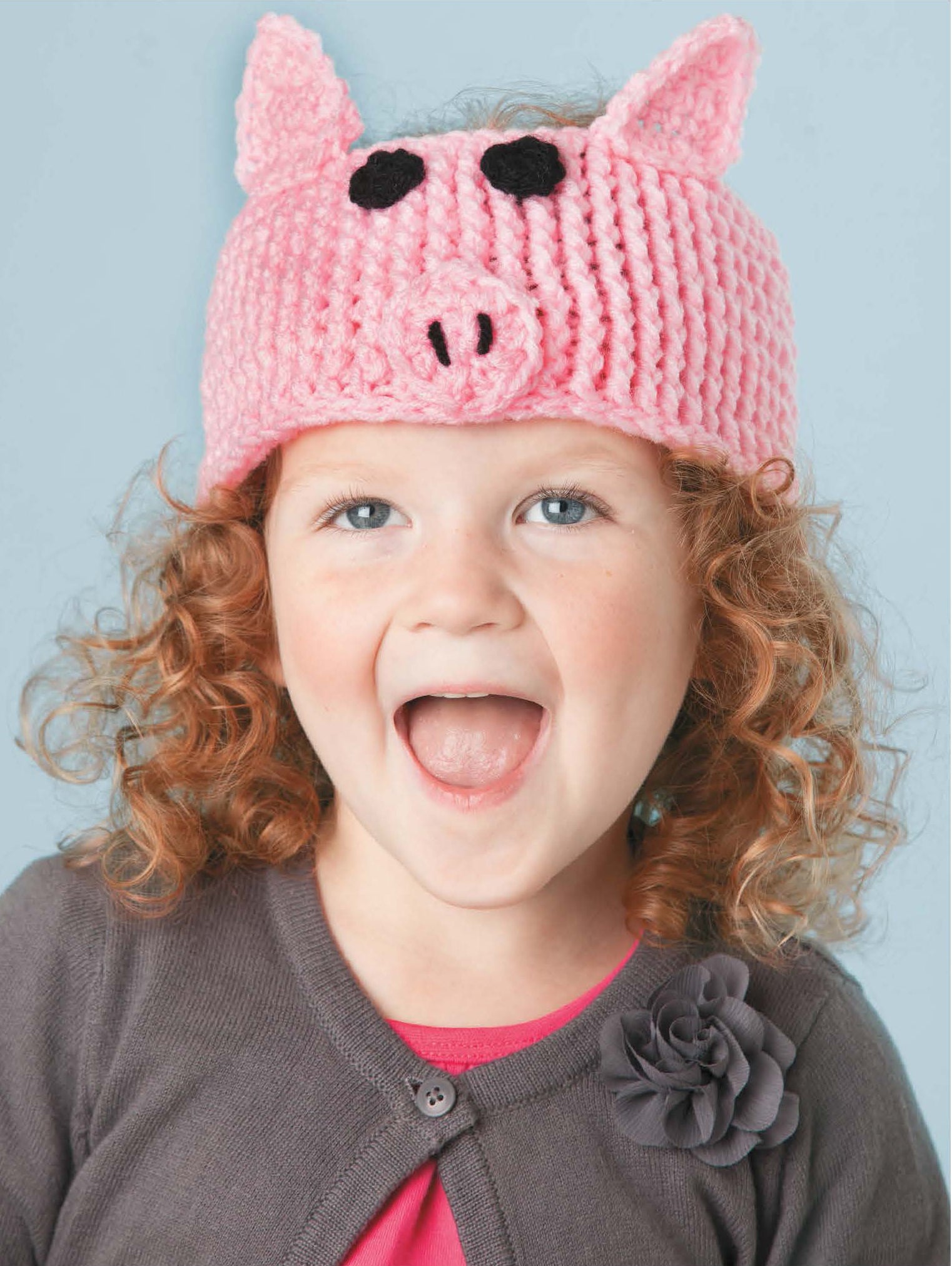 A young child with curly hair smiles widely, wearing Leisure Arts' Little Animals Headgear—a pink knitted baby ear warmer featuring pig ears, eyes, and a snout. She’s dressed in a gray flower cardigan and a pink shirt underneath.