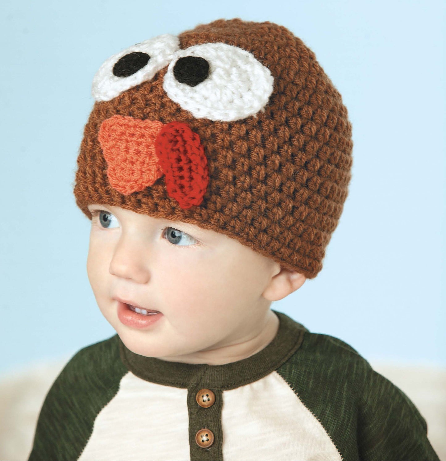 A toddler wears the Leisure Arts "Little Animals Headgear," a brown crochet beanie with big white eyes, red beak, and wattle like a turkey, paired with a green and white shirt against a light blue background.