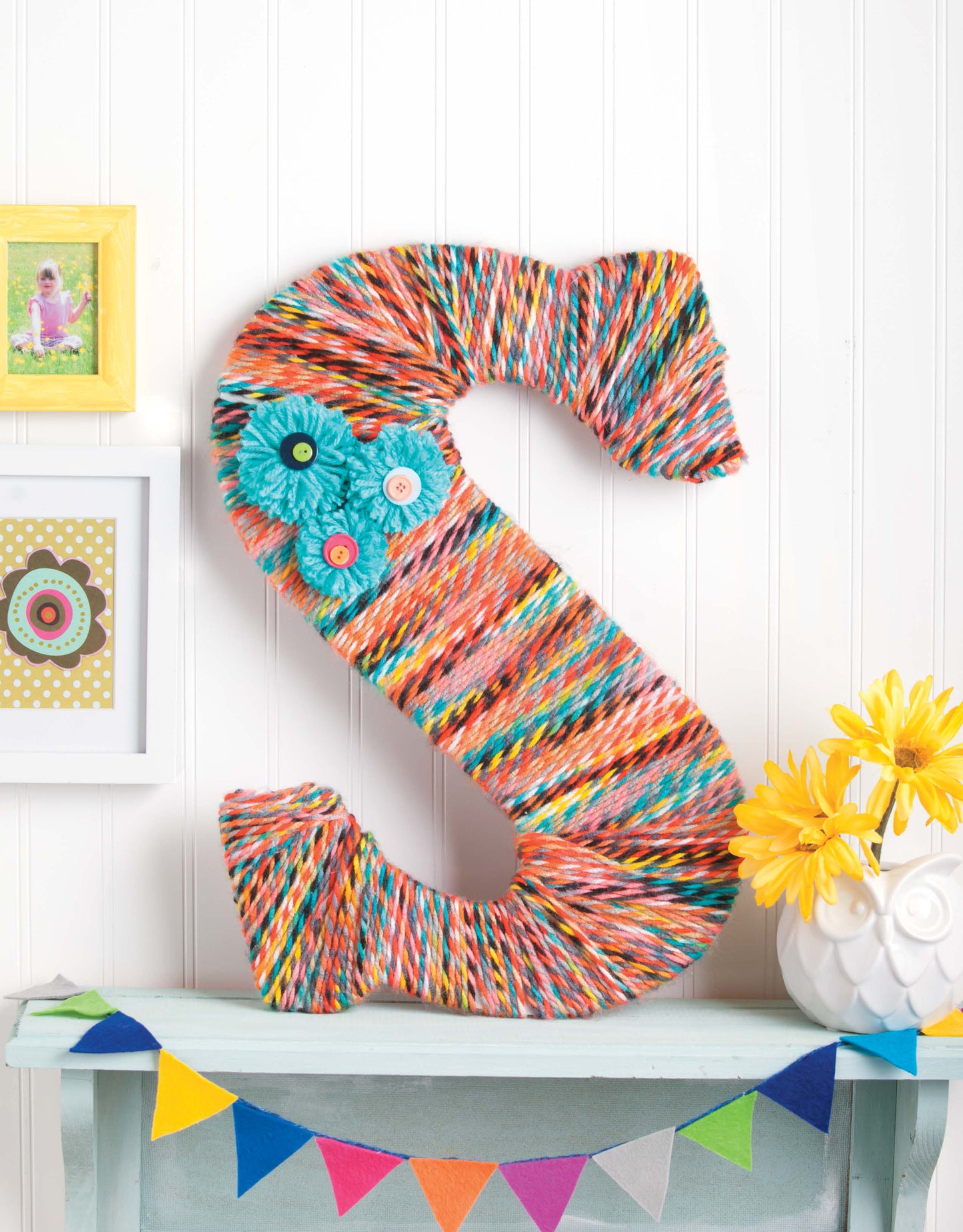A Decorating Letters Digital Download by Leisure Arts featuring a large letter S wrapped in colorful yarn with felt flowers is displayed on a blue shelf, alongside an owl vase with yellow flowers, framed photos, and bunting for a charming decor look.
