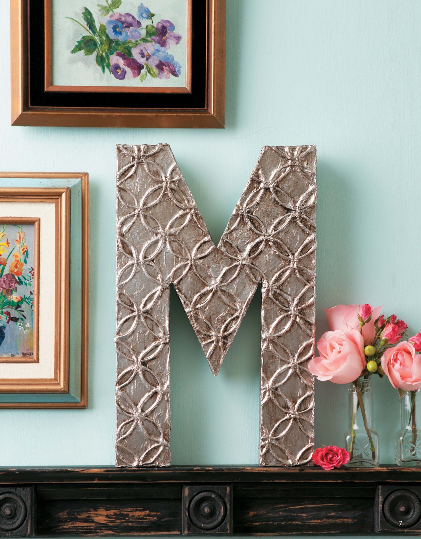 A unique M from the Leisure Arts Decorating Letters Digital Download features a metallic geometric design on a dark shelf, paired with pink roses and floral paintings, creating charming home décor against a pale blue wall.