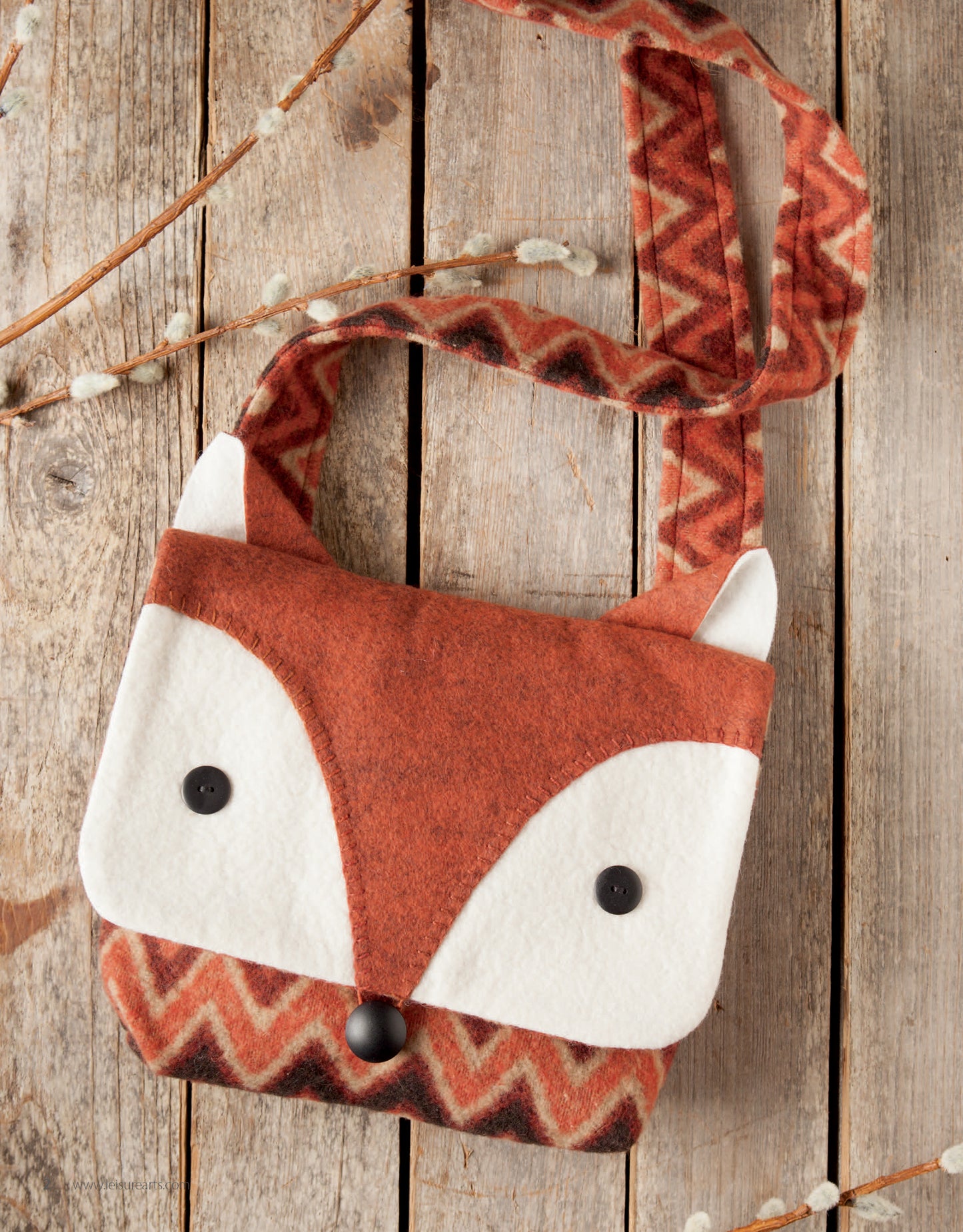 The Felt Crafty Critters Digital Download by Leisure Arts features an easy-to-sew fox face purse with white accents, button eyes, and a patterned orange strap, displayed on rustic wood beside pussy willow branches.