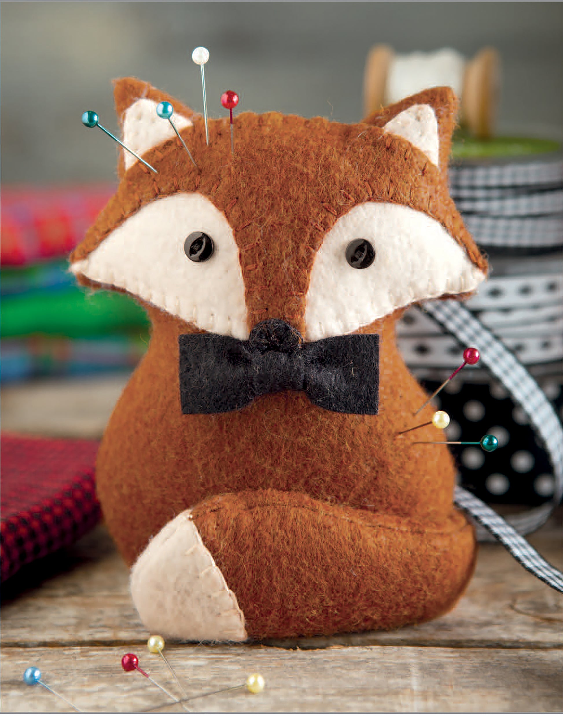 The Felt Crafty Critters Digital Download by Leisure Arts features a fox-shaped wool felt pincushion with a black bow tie and colorful pins—an easy embroidery project with blurred fabrics and ribbon spools in the background.