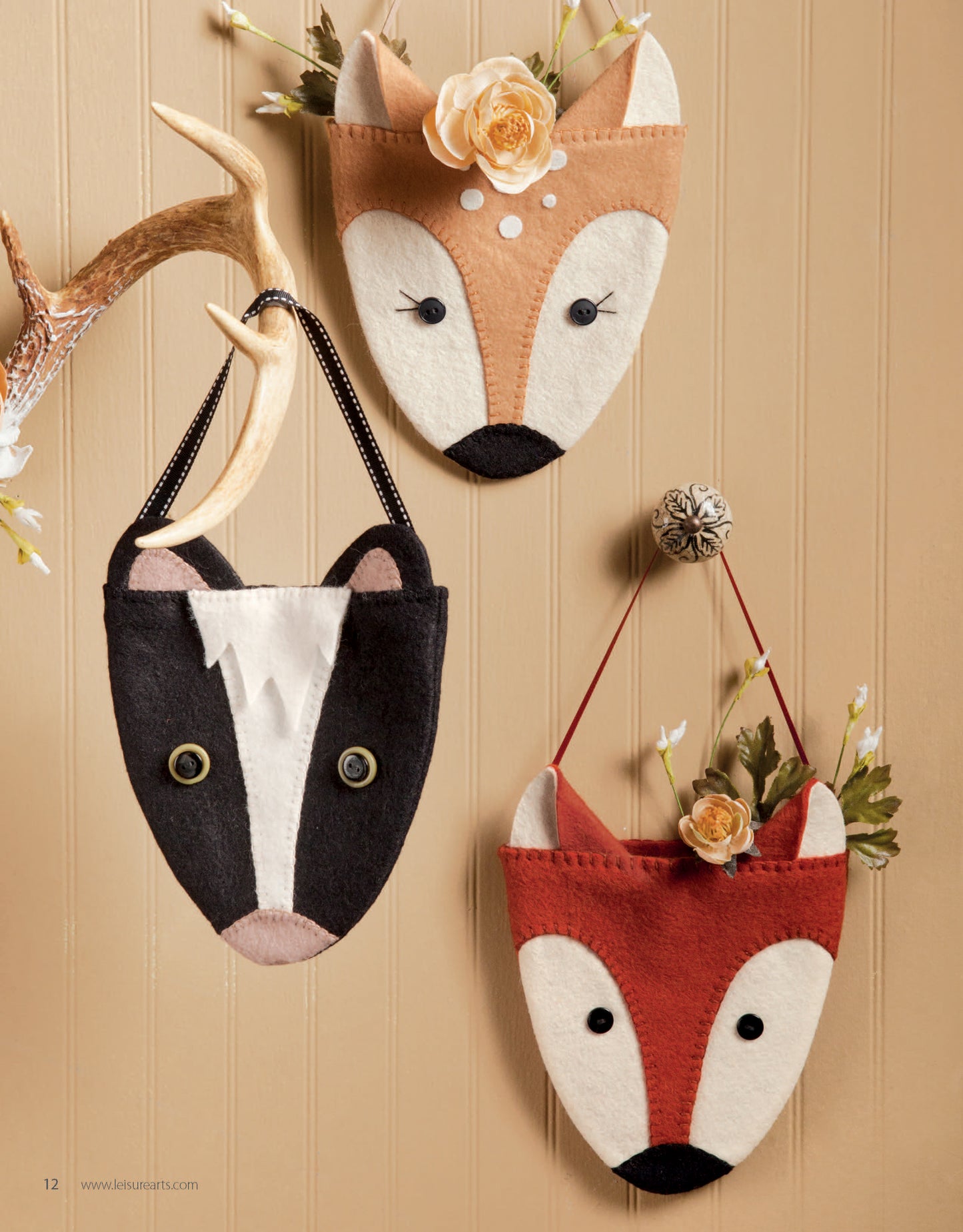 Displayed on a tan wall, the deer and skunk pouches from Leisure Arts' Felt Crafty Critters Digital Download are easy-to-sew organizers, perfect for wool felt pincushions, and hang from an antler-shaped hook with decorative flowers.