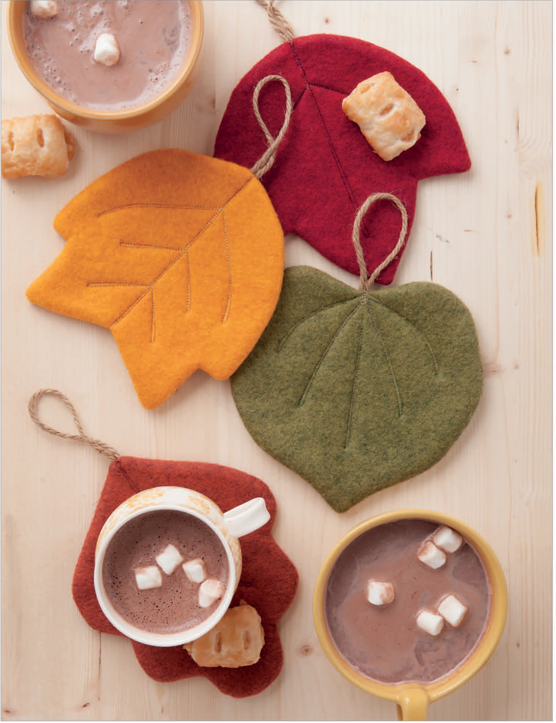 Three mugs of hot chocolate with marshmallows sit on a table with puff pastries, leaf-shaped coasters in autumn colors, and pincushions made using the Felt Crafty Critters Digital Download from Leisure Arts for cozy charm.