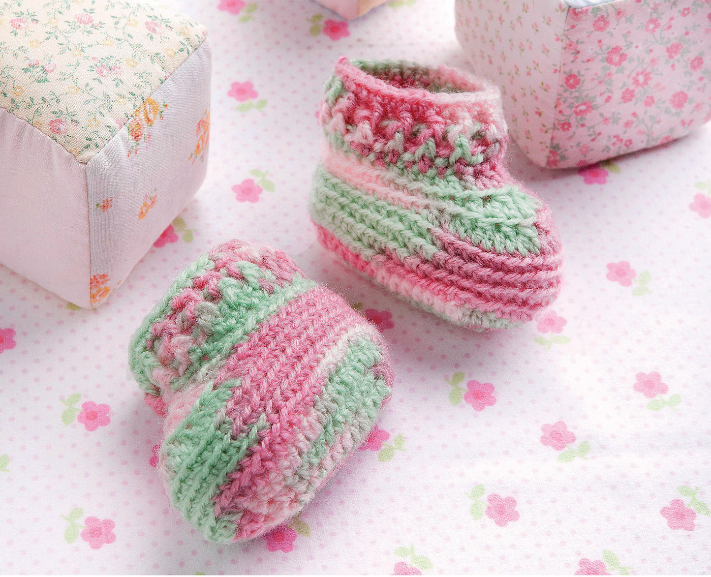 Two pastel pink, green, and white baby booties rest on a floral blanket with fabric blocks—an adorable gift idea or knitting inspiration from "One-Skein Baby Projects" by Leisure Arts.