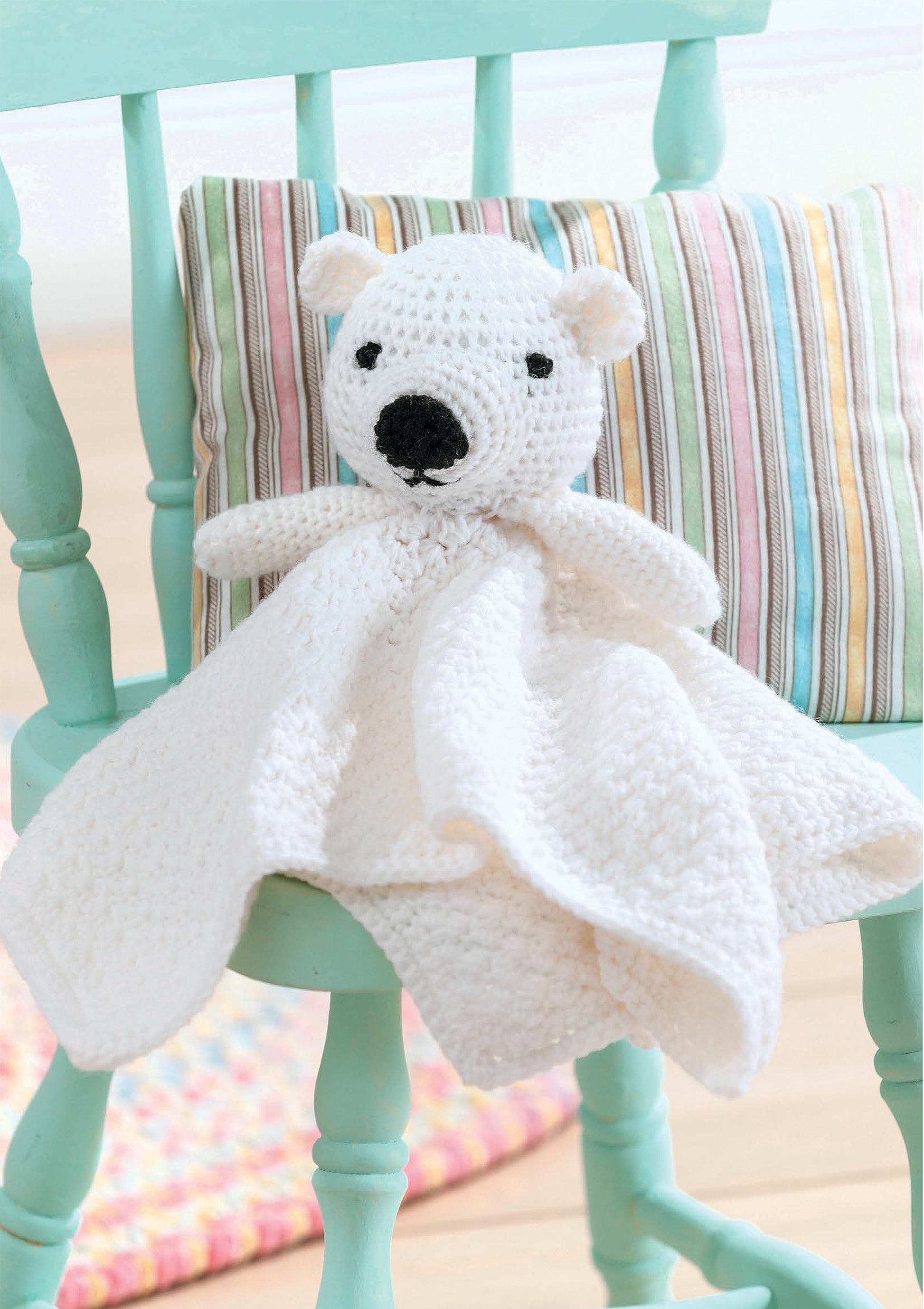 The Leisure Arts One-Skein Baby Projects features a crocheted white bear comforter with a black nose and a soft, blanket-like body—ideal as a newborn gift. Shown on a pastel-striped pillow atop a light turquoise wooden chair.