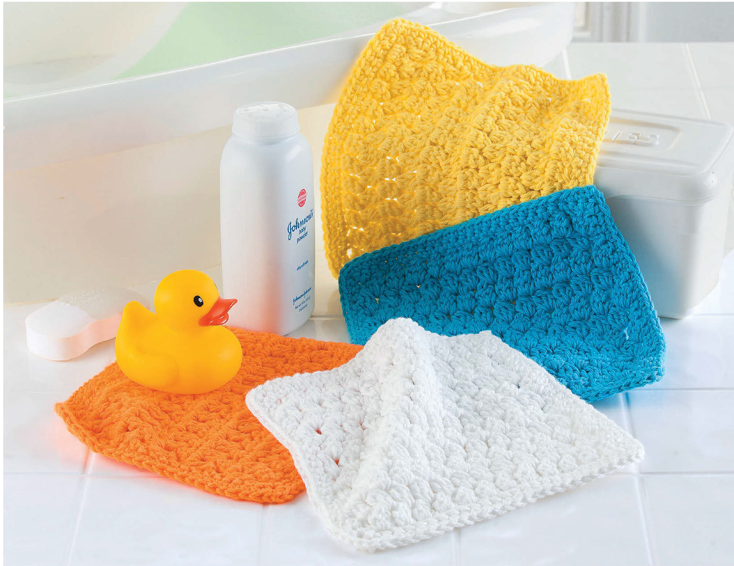 Four crocheted washcloths in yellow, blue, orange, and white are arranged near a bathtub with a rubber duck and baby powder. From One-Skein Baby Projects by Leisure Arts—perfect gifts for newborns or quick crochet projects.